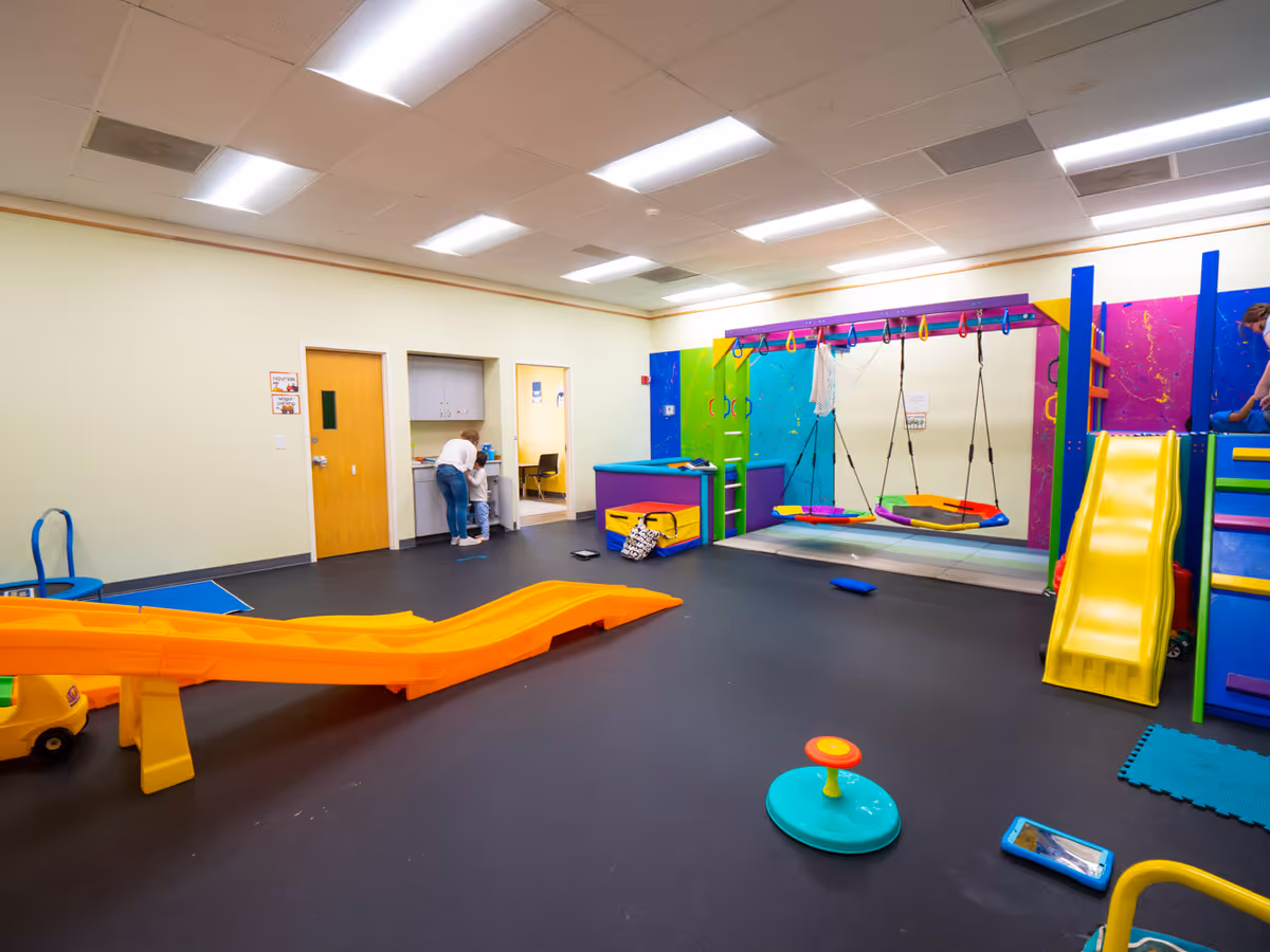 Colorful indoor play area with swings and slides, adults near a kitchenette in background.