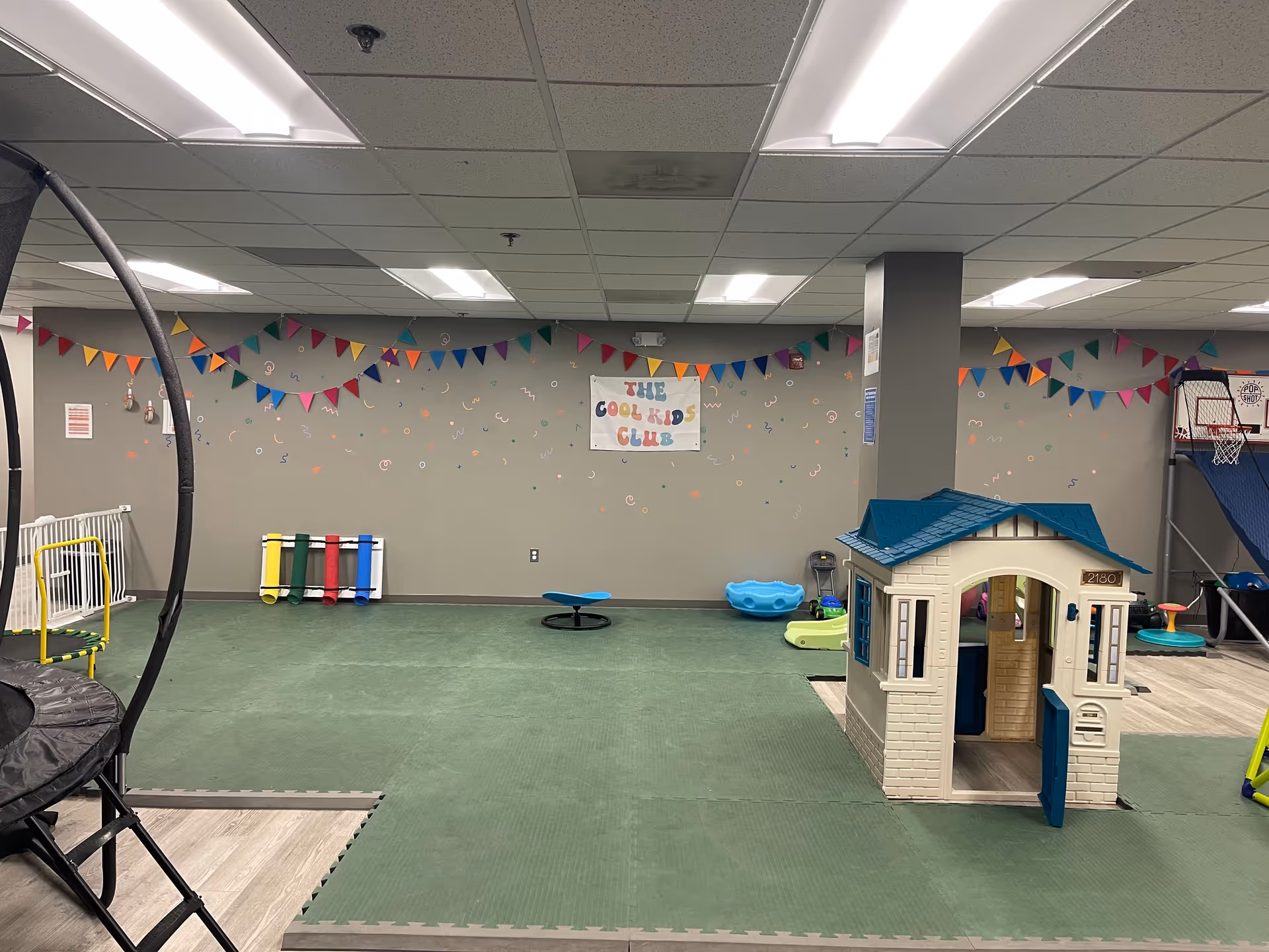 Indoor children's playroom with a plastic house, mats, and colorful banners.