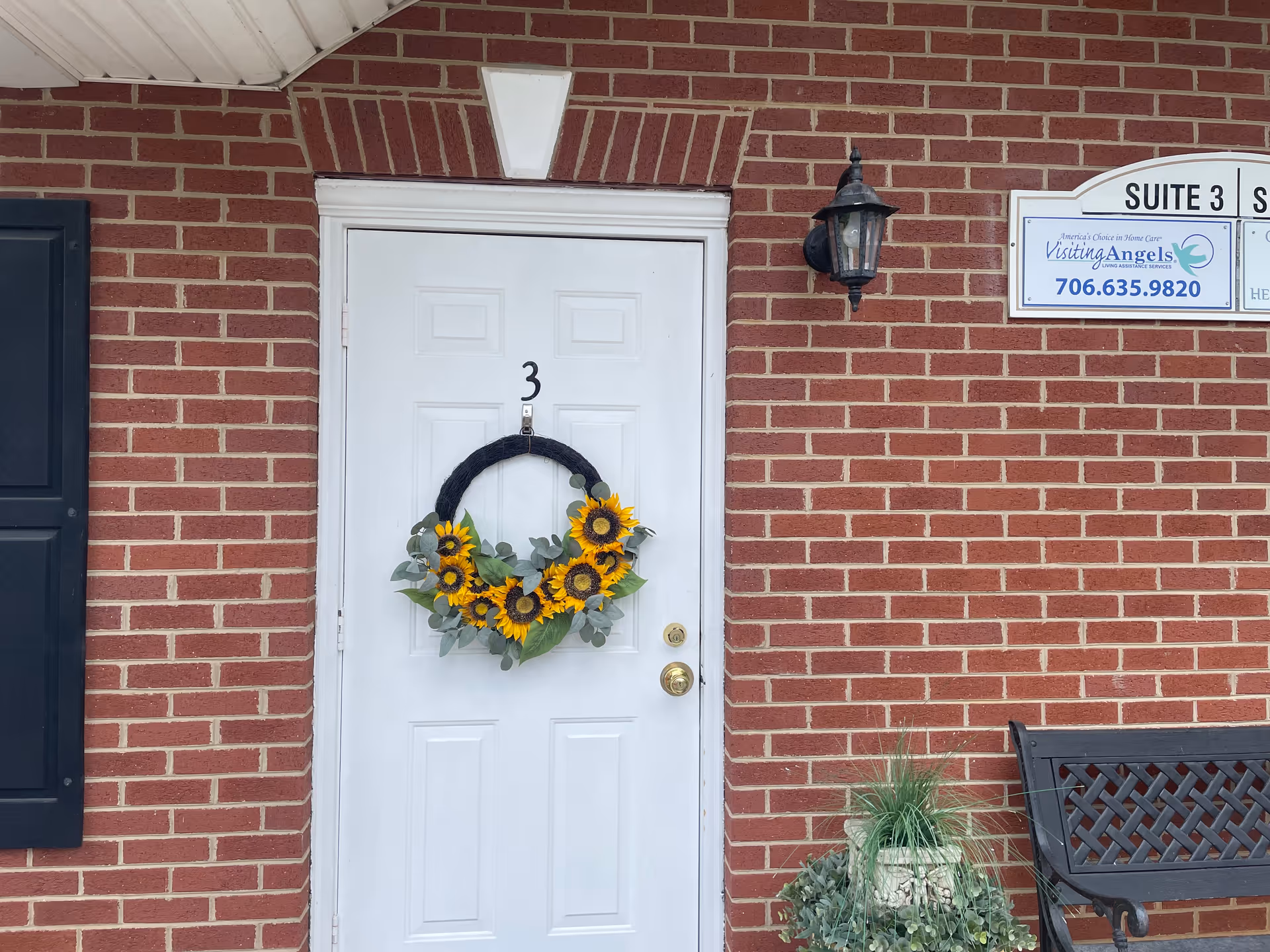 White front door with sunflower wreath on brick building exterior, beside bench and Visiting Angels sign.