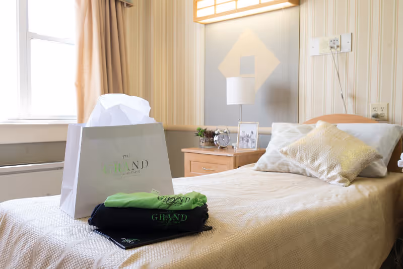 A neatly made bedroom in a senior living facility with bed, nightstand, lamp, and bag.