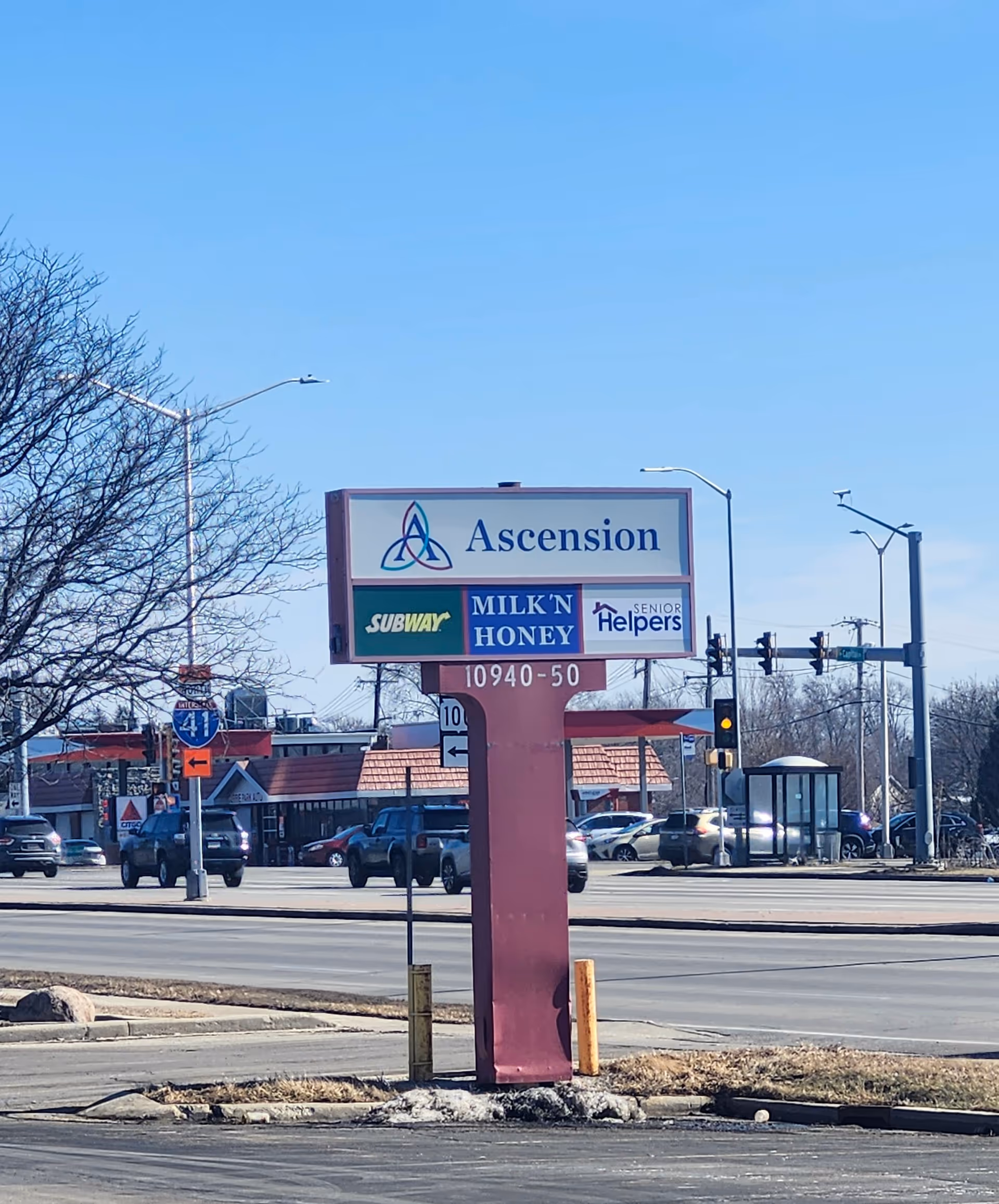 Street-level exterior with a tall pink sign advertising Ascension and Senior Helpers beside traffic.