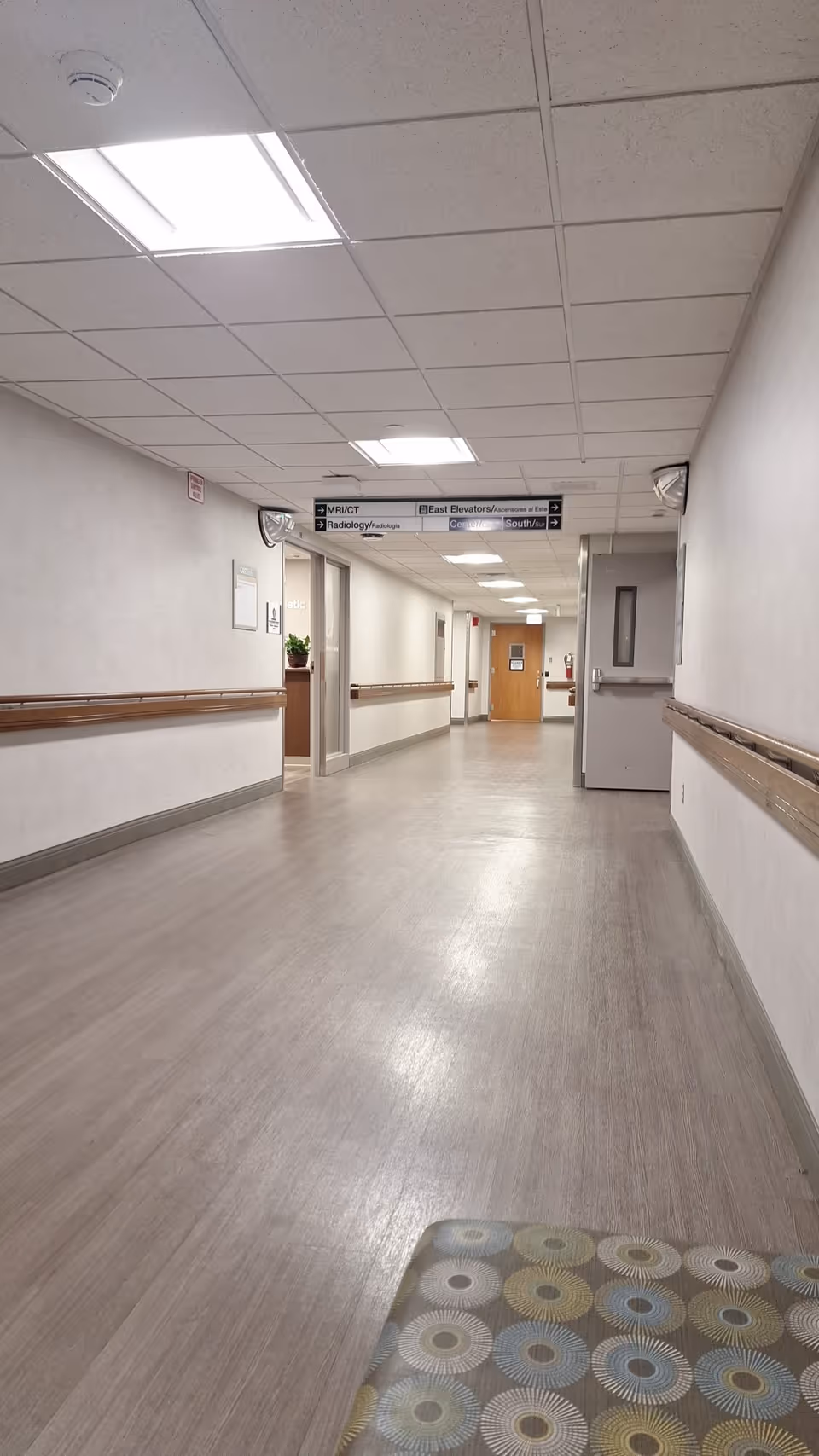 Long hospital corridor with ceiling lights, white walls, handrails, and doors along the hall.