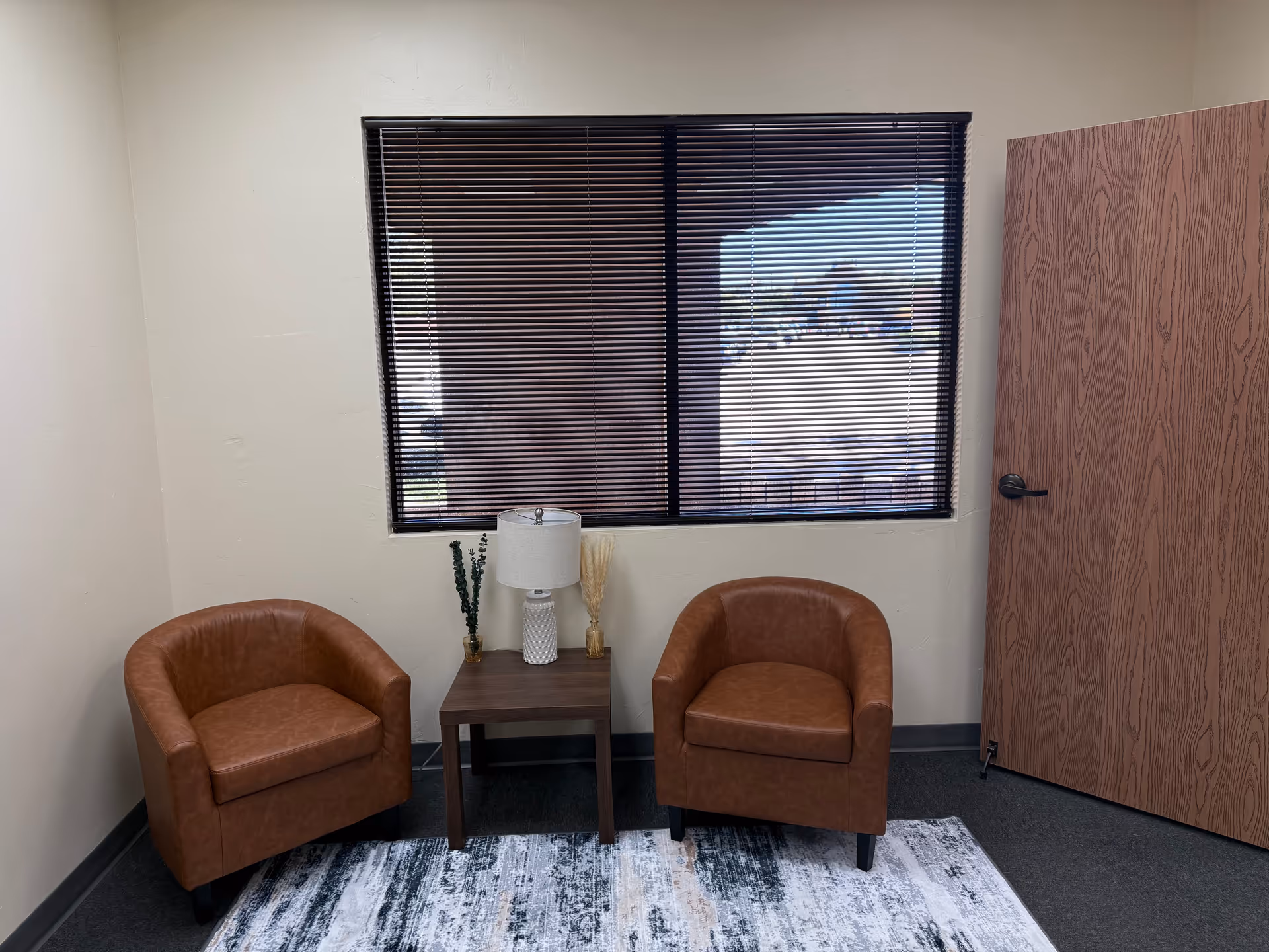 Two brown armchairs flank a table with a lamp beneath a window with blinds.
