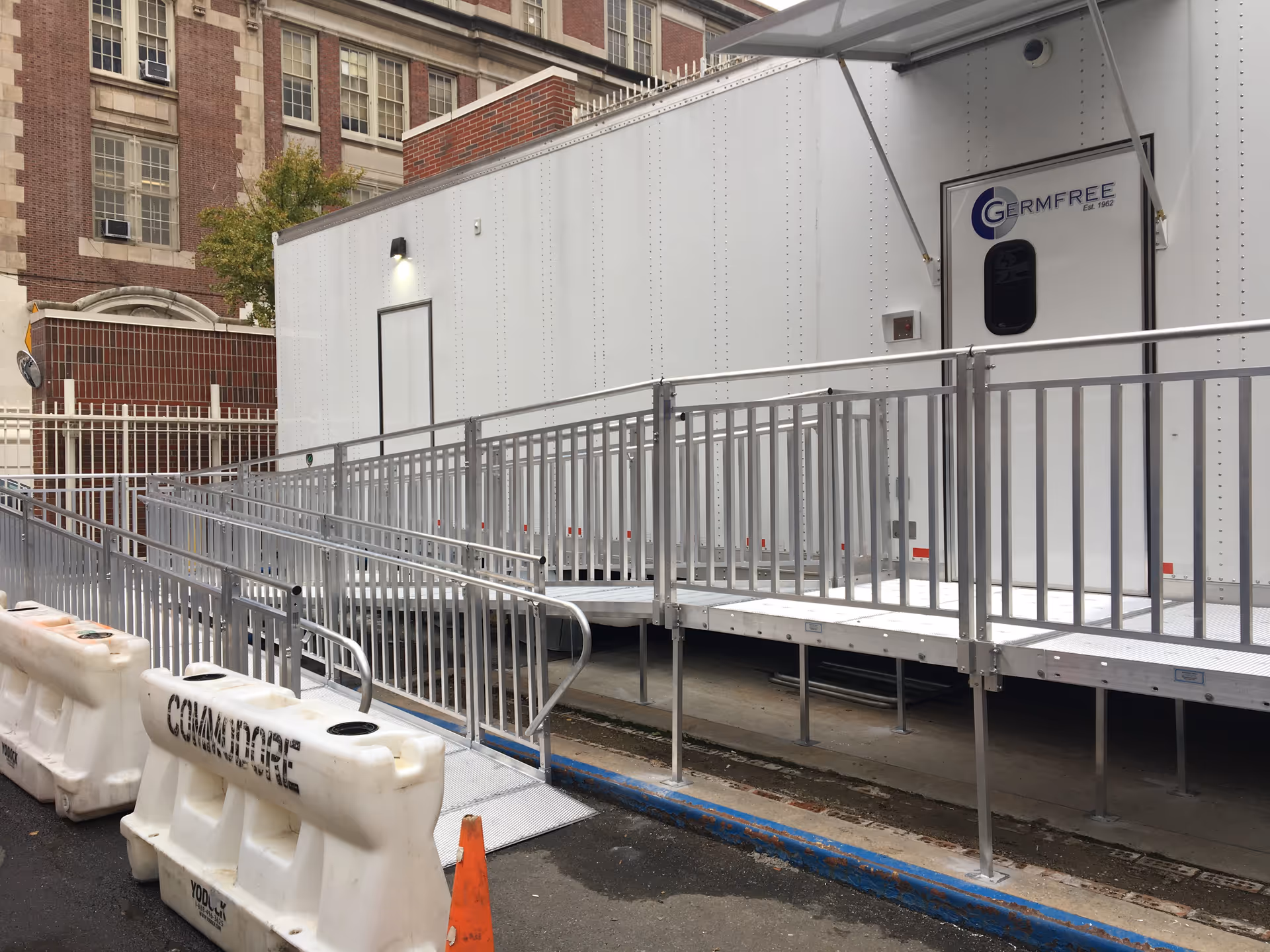 Exterior view of a white modular trailer with ramps and railing beside a brick building.