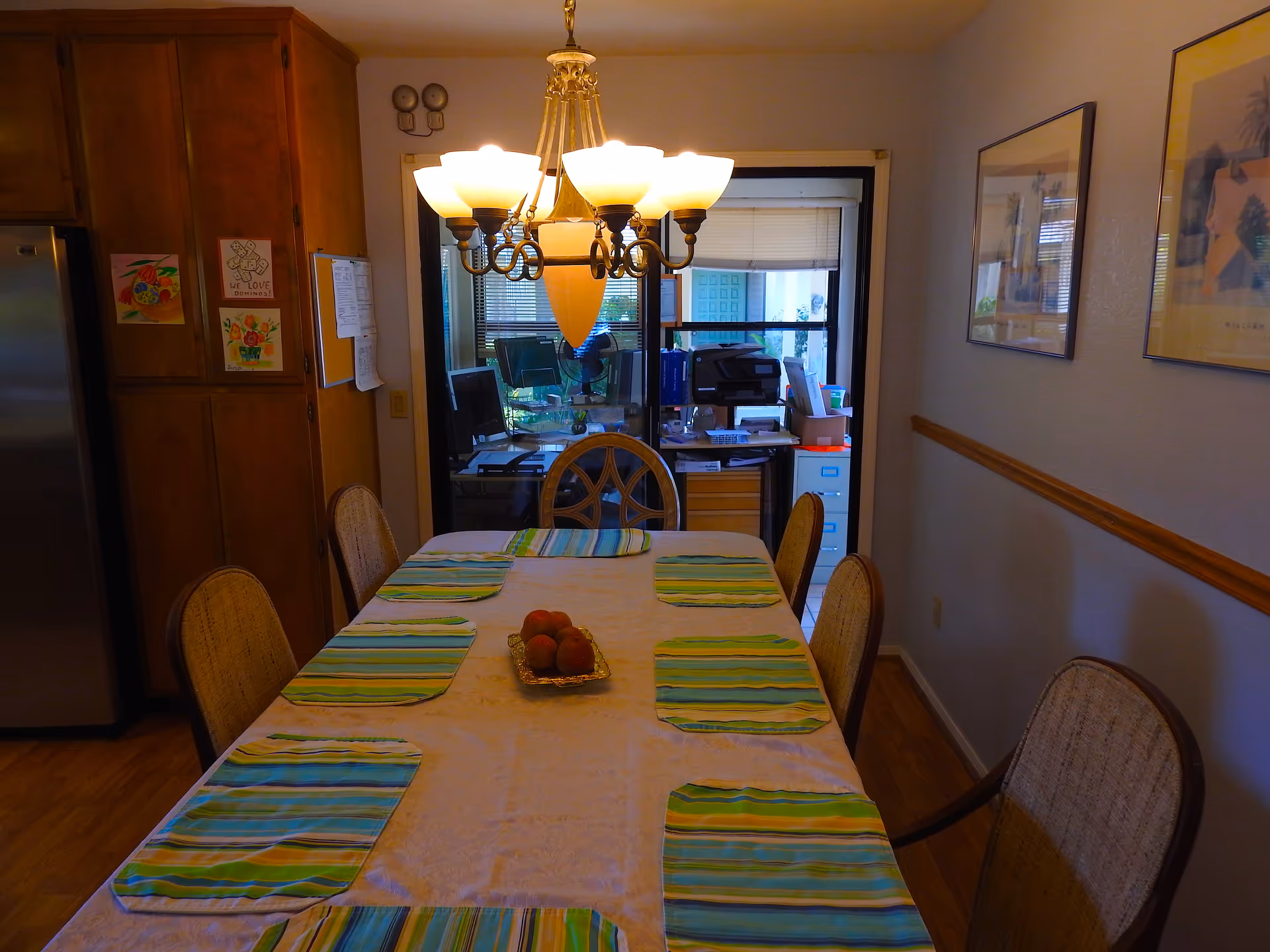 Dining room table with striped placemats and a chandelier overlooking a kitchen.