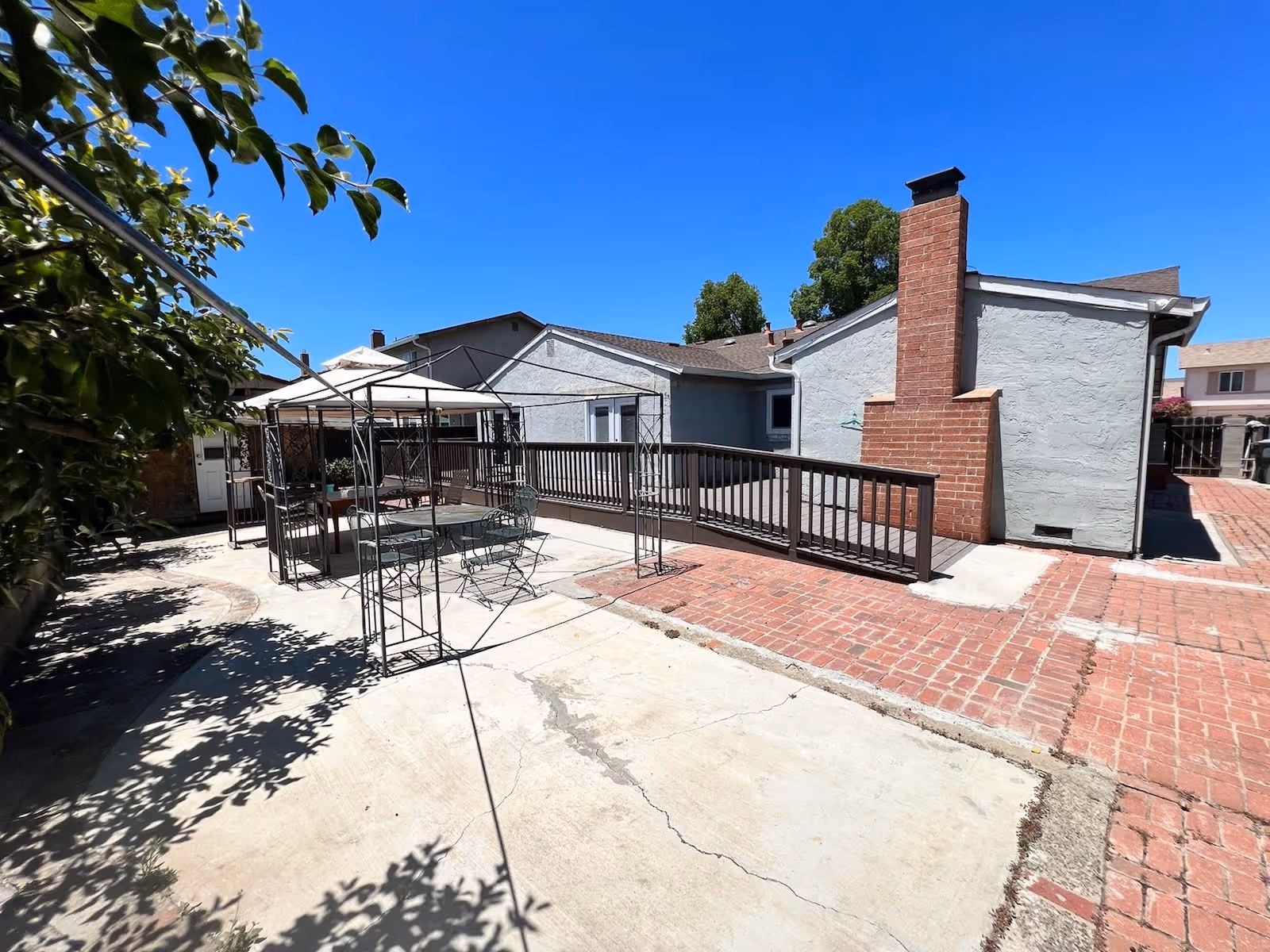 Sunny outdoor courtyard with metal gazebo frames, brick paths, and seating beside a building.