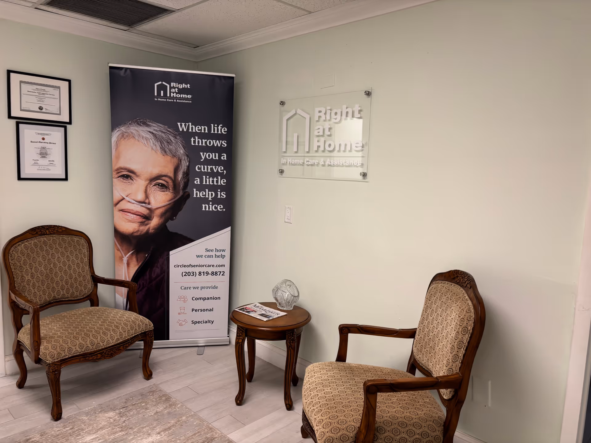 Two upholstered chairs, small round table, and a Right at Home banner in a reception area.