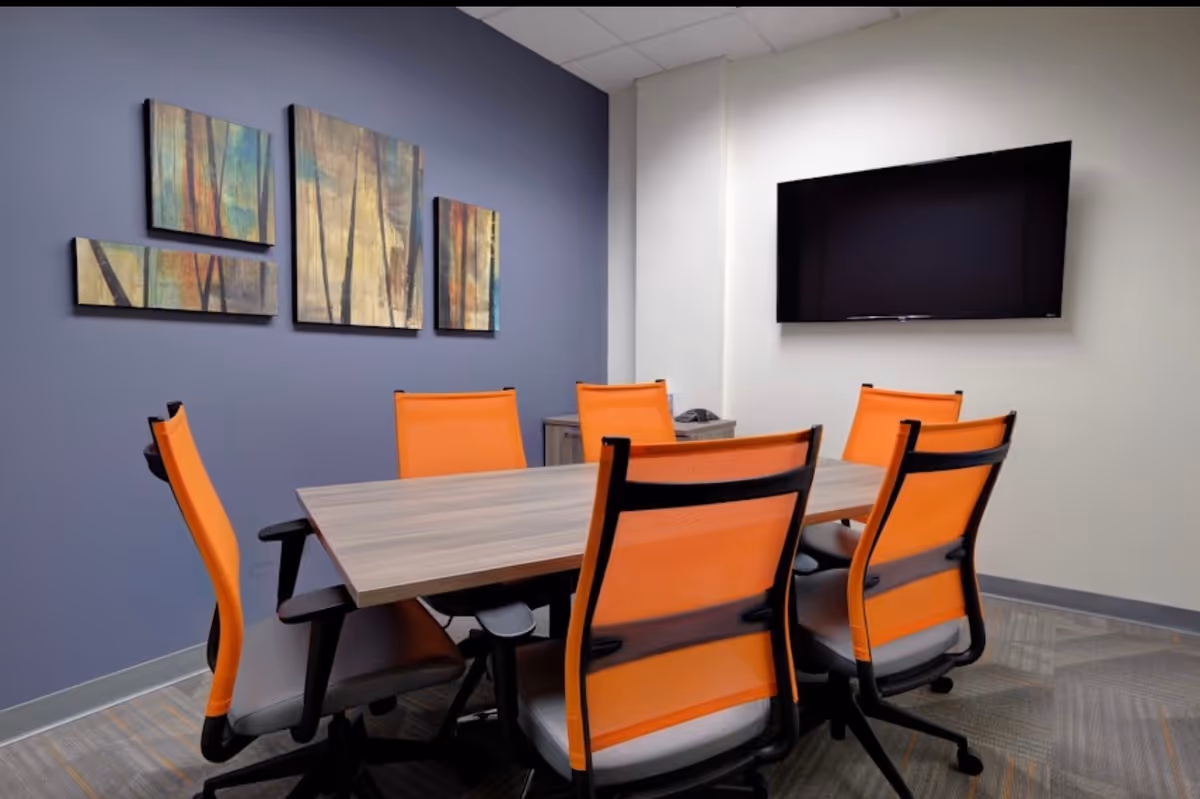 Meeting room with a wooden table, orange chairs, wall art, and a wall-mounted TV.