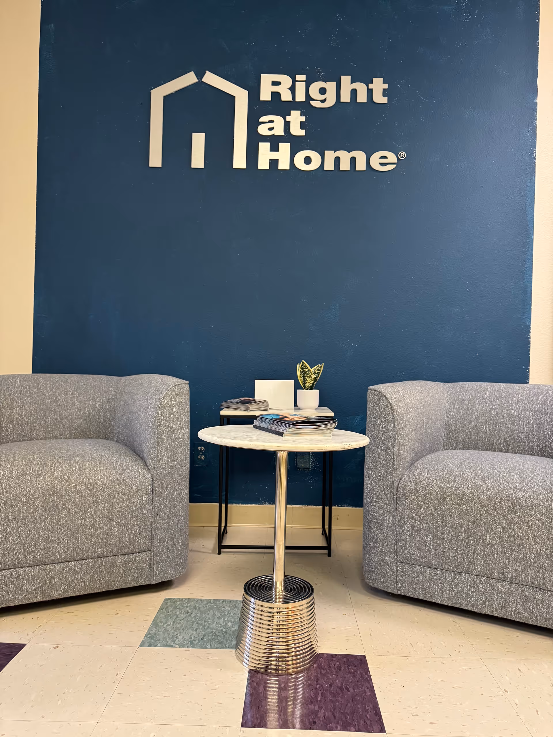 Blue wall with Right at Home logo flanked by two gray lounge chairs and a table