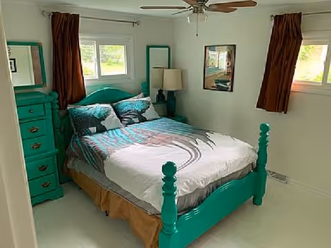Turquoise bed frame and dresser in a small bright bedroom with windows and brown curtains.