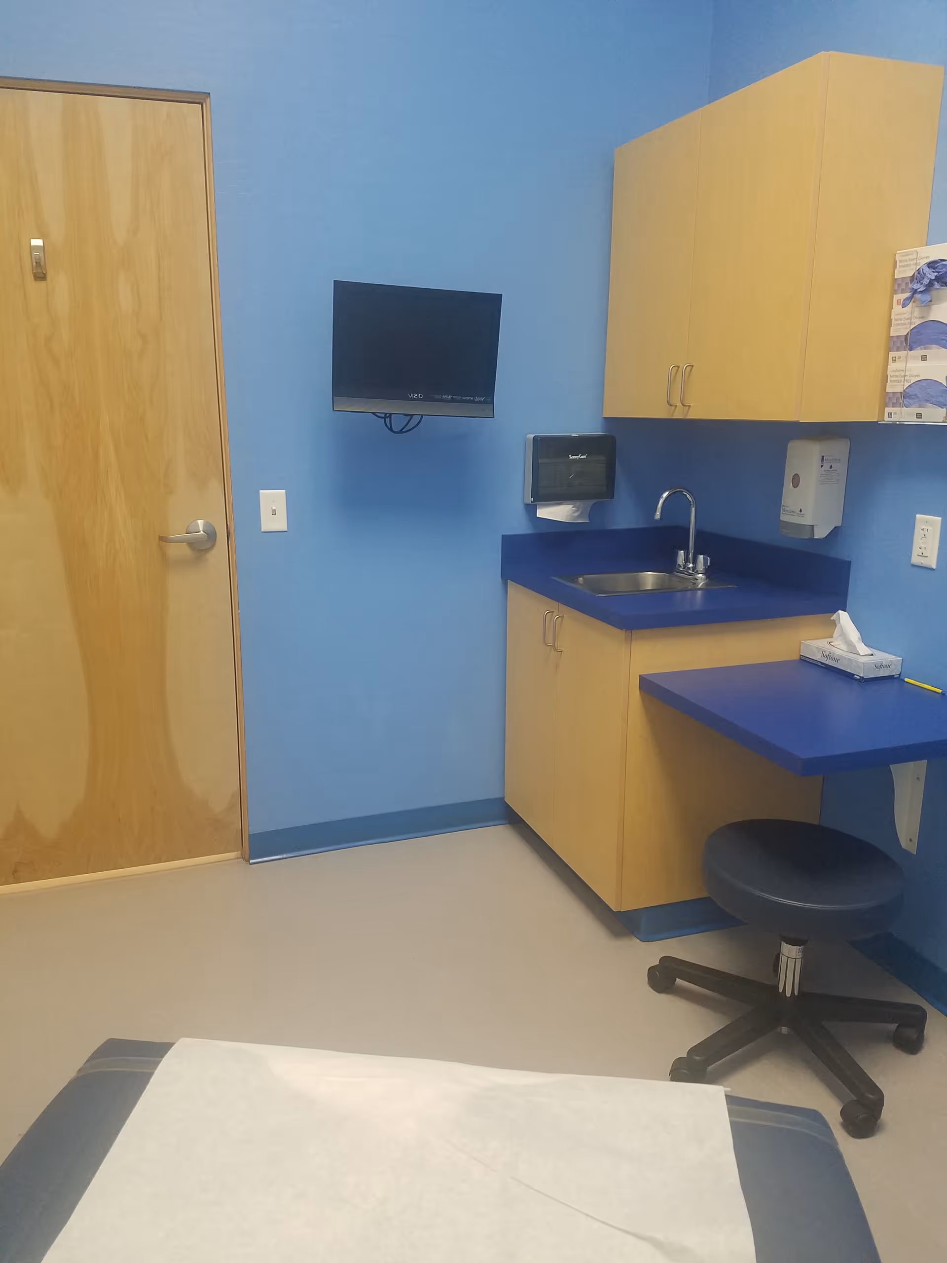 Blue-walled senior living room kitchenette with sink, cabinets, tissue dispenser, and wall-mounted TV.