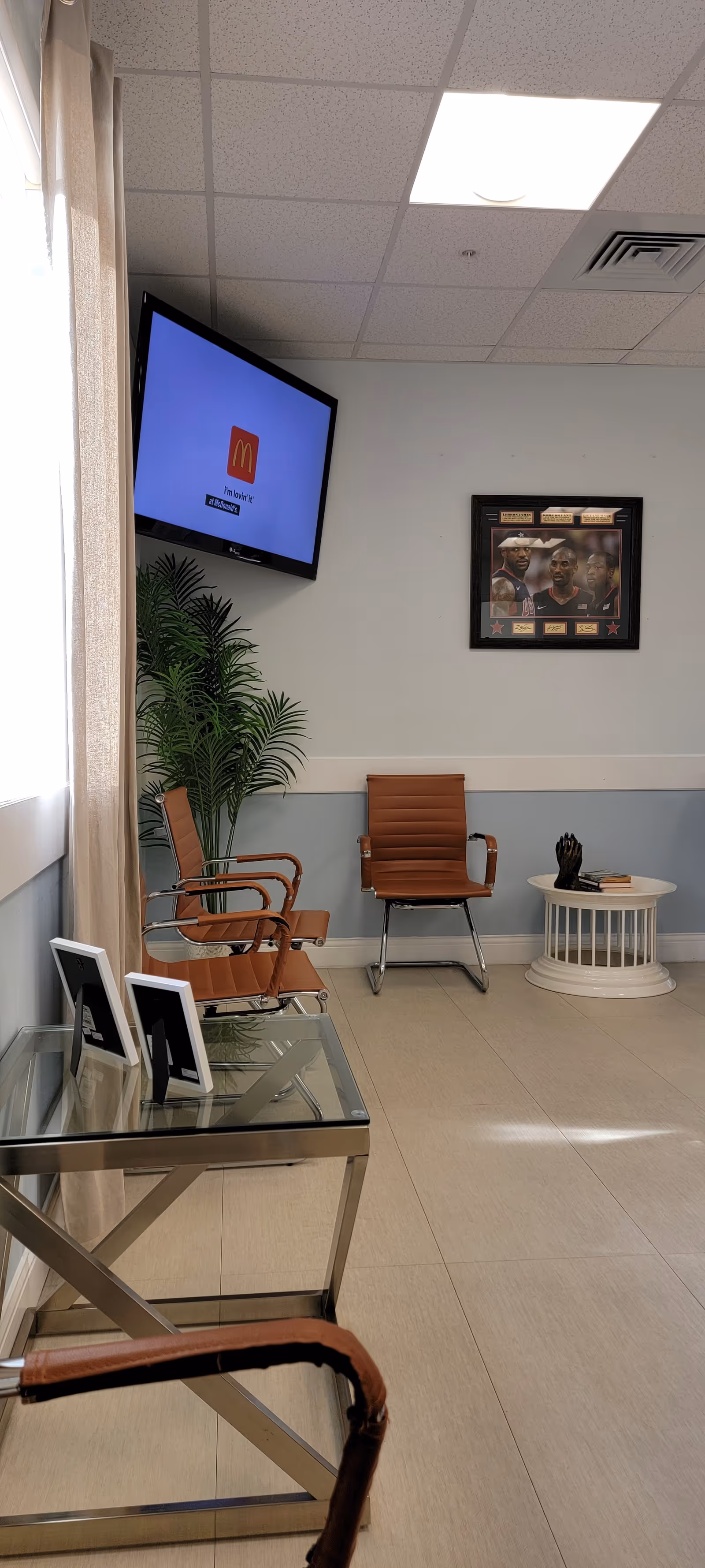 Small hospital waiting area with brown chairs, a mounted TV, plant, and glass table.