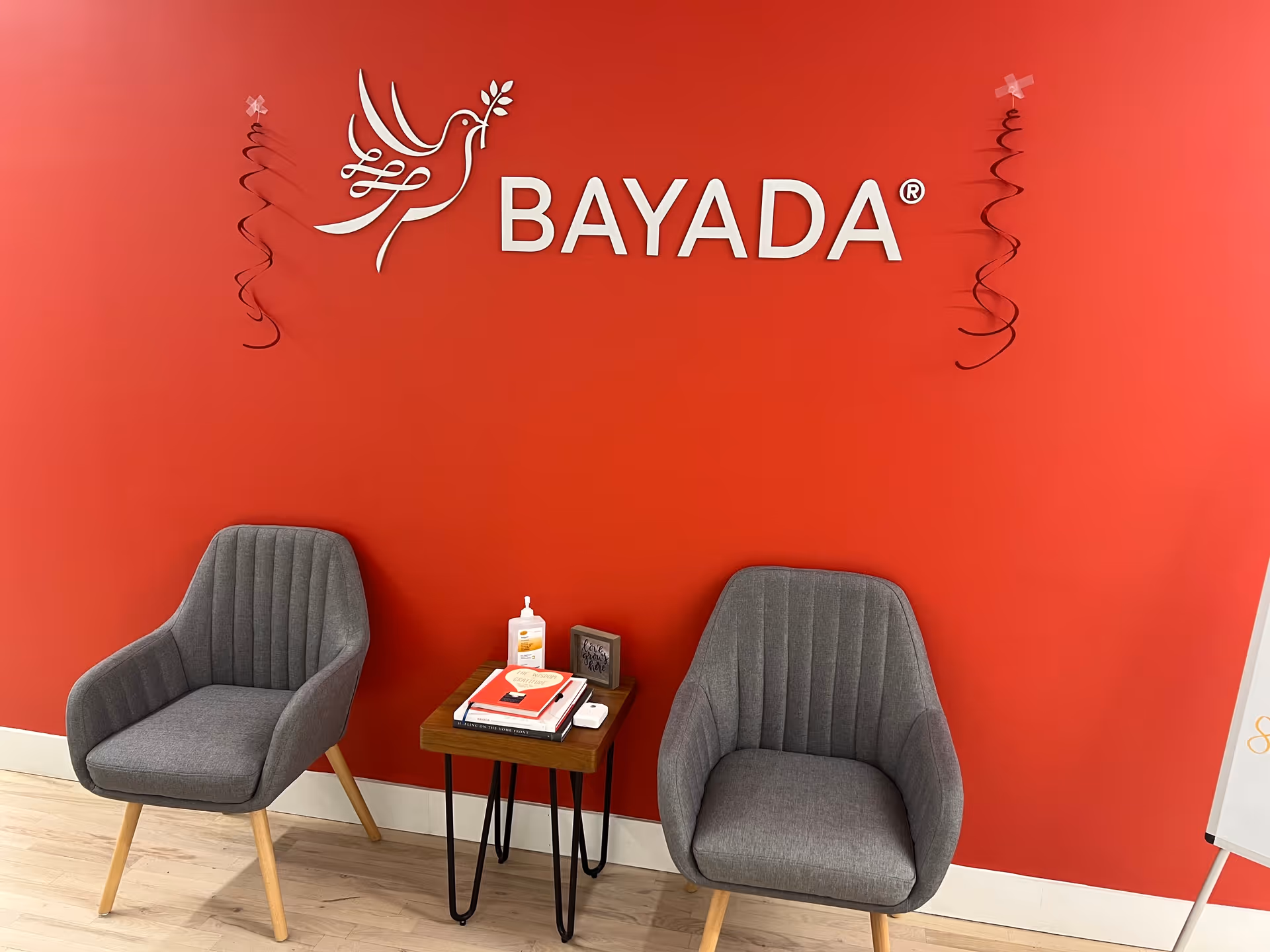 Two gray armchairs sit beside a small table against a red wall with BAYADA logo.