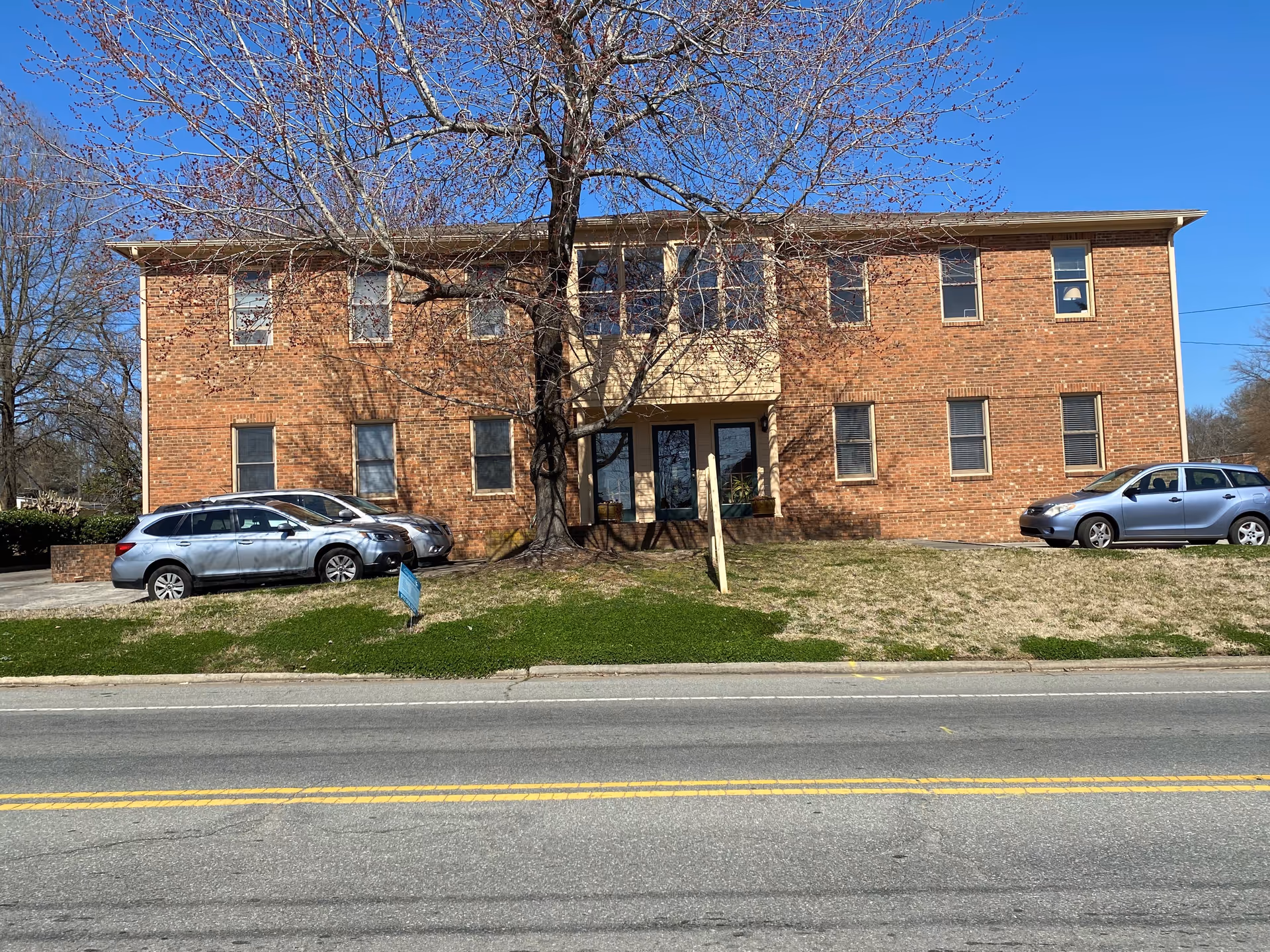 Brick two-story senior living building with a leafless tree and parked cars.