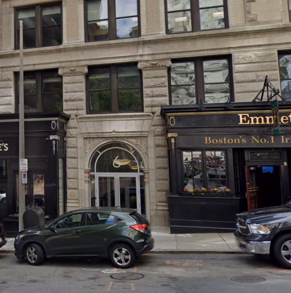 Sandstone building facade with arched doorway and storefront windows, parked cars on the street.
