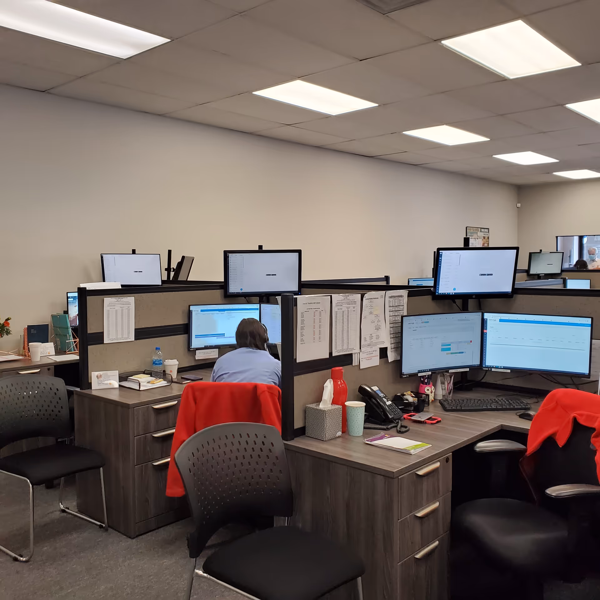 Office cubicles with multiple computer monitors and a staff member at a desk.