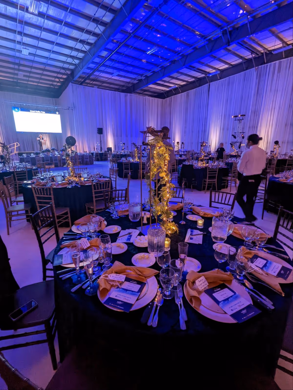 Indoor banquet hall with round tables, gold centerpieces, and blue lighting.