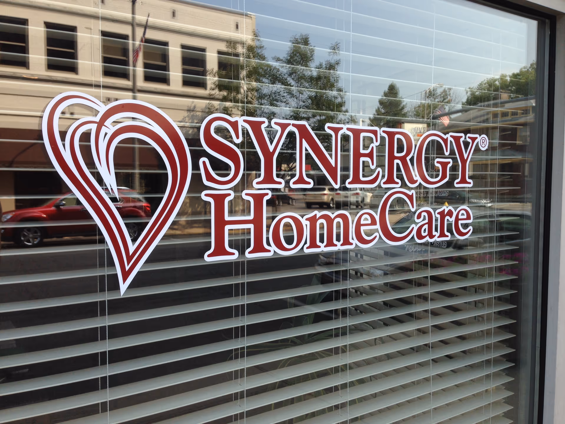 Storefront window displays SYNERGY HomeCare sign with heart logo and blinds.