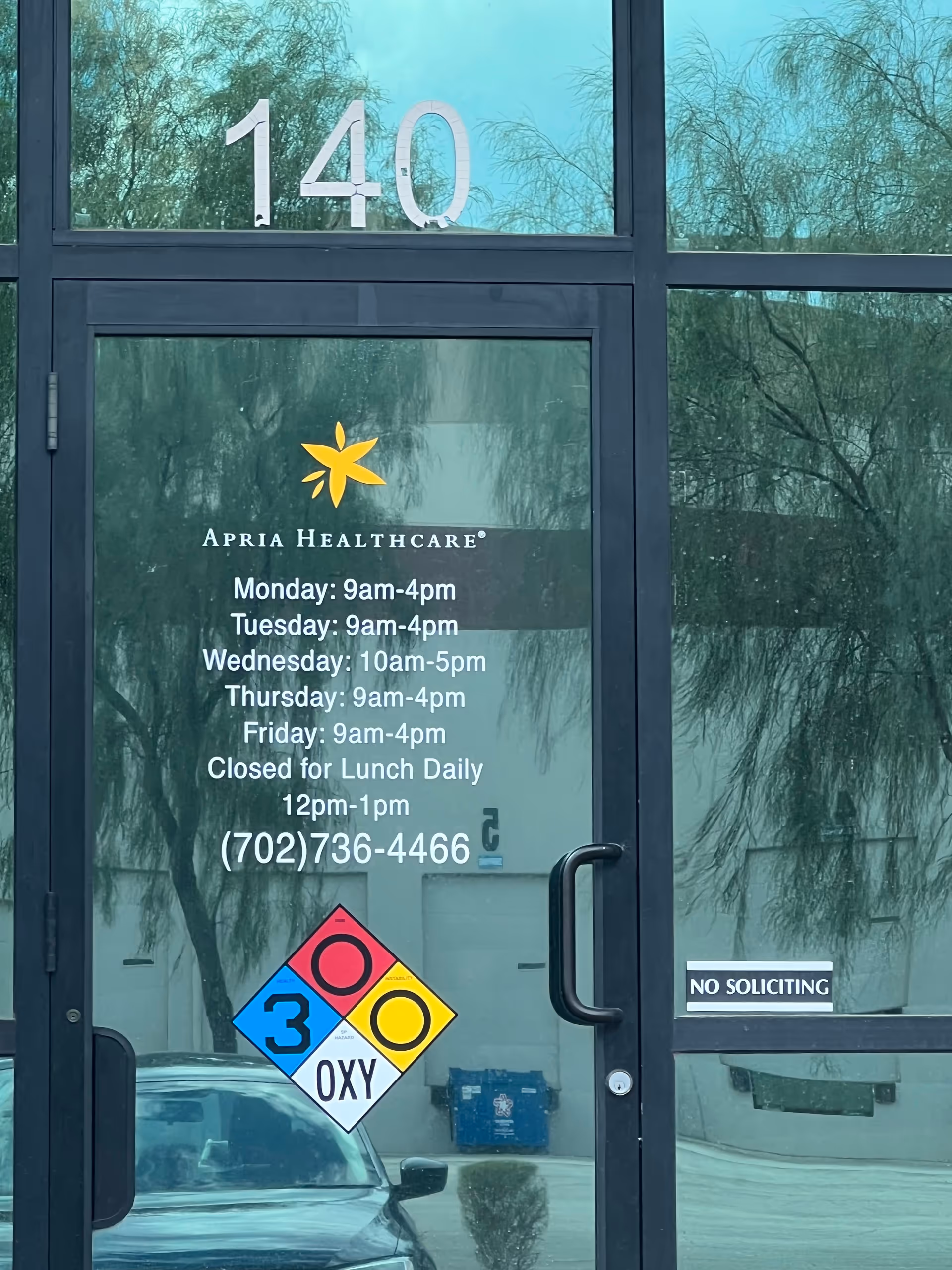 Glass front entrance of Apria Healthcare displaying hours and a No Soliciting sign