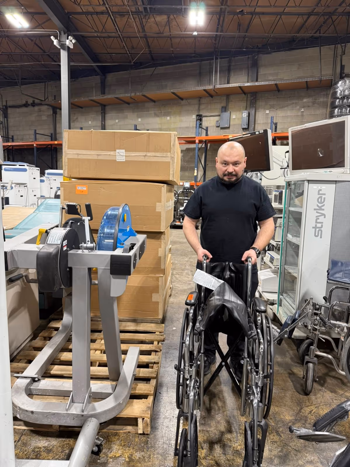 A man in a wheelchair stands in a cluttered indoor warehouse with pallets and equipment.