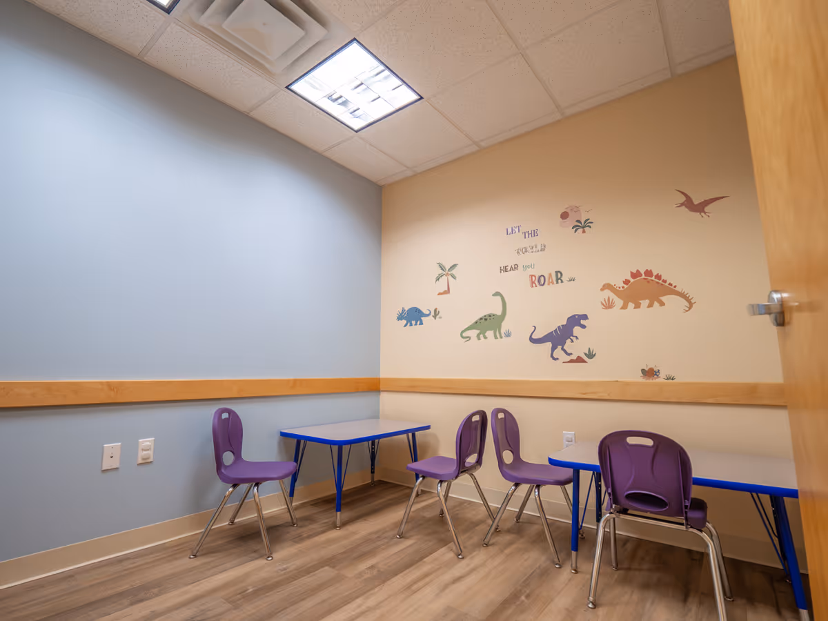 Small activity room in a senior facility with purple chairs, blue tables, and dinosaur wall art.