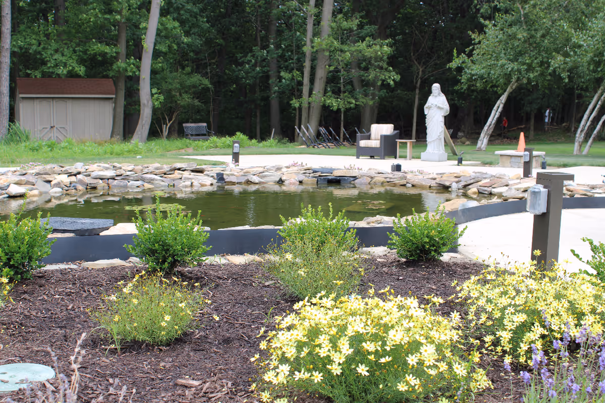 A serene outdoor garden with a pond, statue, patio seating, and blooming shrubs.