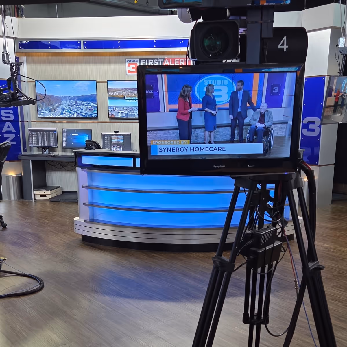 Television studio with a tripod-mounted monitor showing Synergy HomeCare on-air graphics.