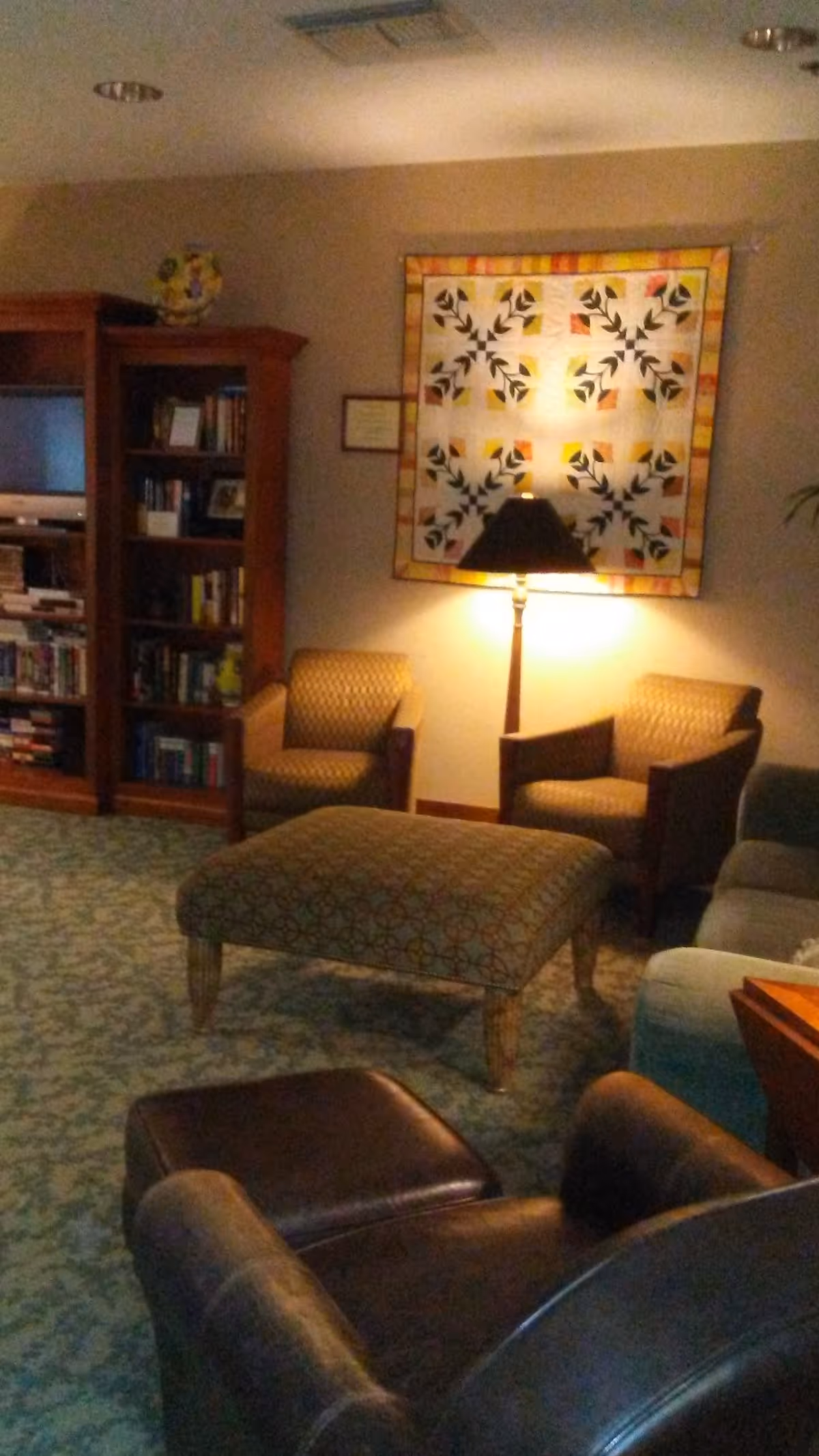 Cozy lounge area with armchairs, ottoman, bookshelf, lamp, and quilted wall hanging.