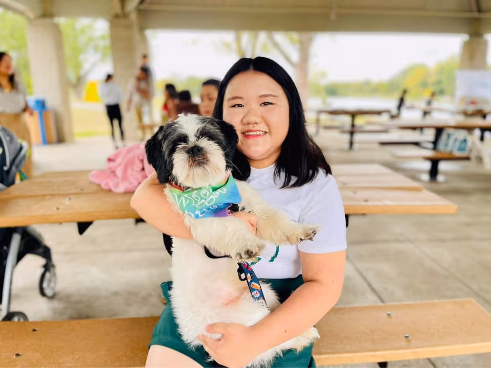 A smiling woman holds a fluffy dog at an outdoor picnic pavilion.