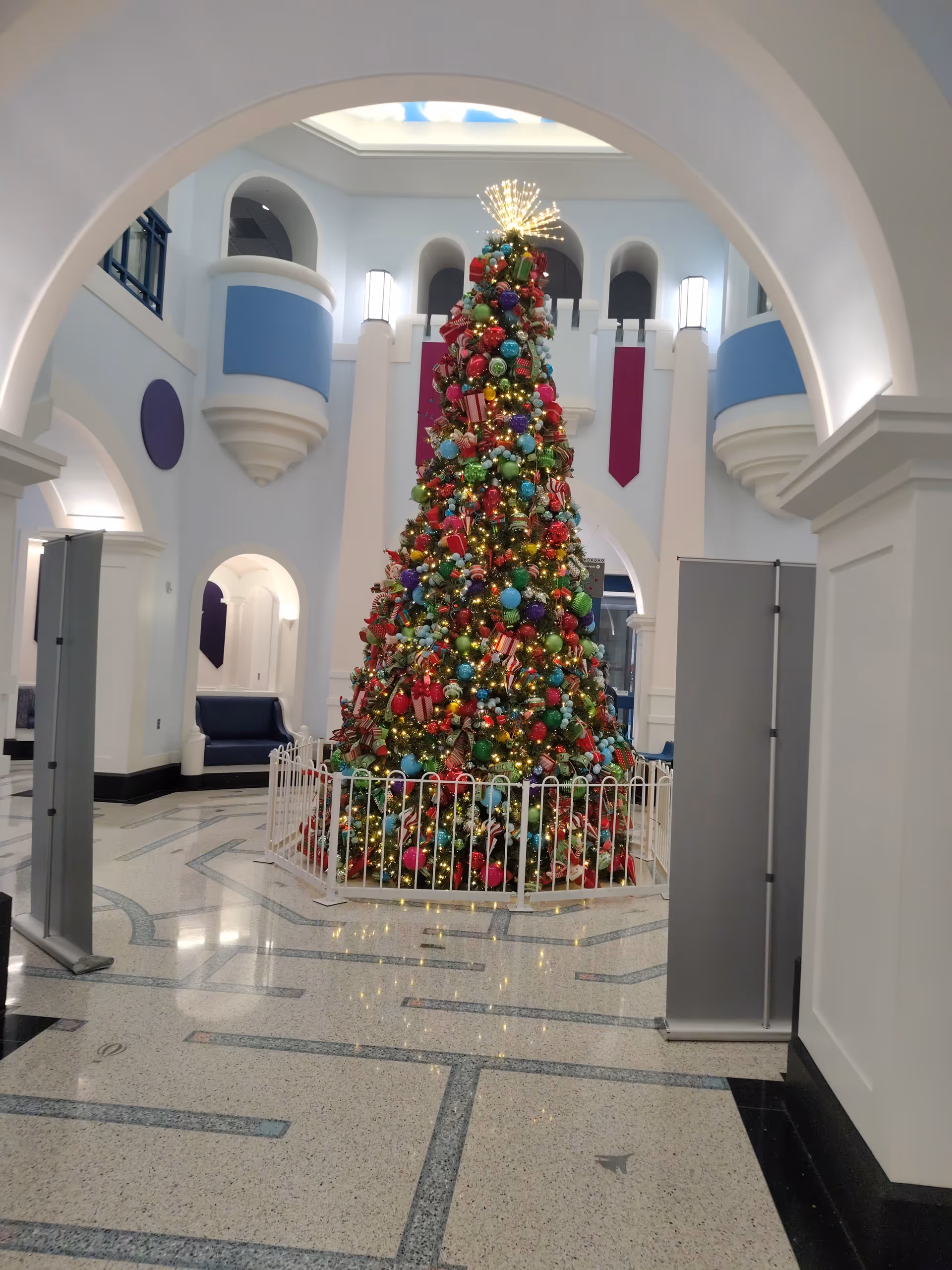 Ornate indoor lobby with a tall decorated Christmas tree enclosed by a white fence.