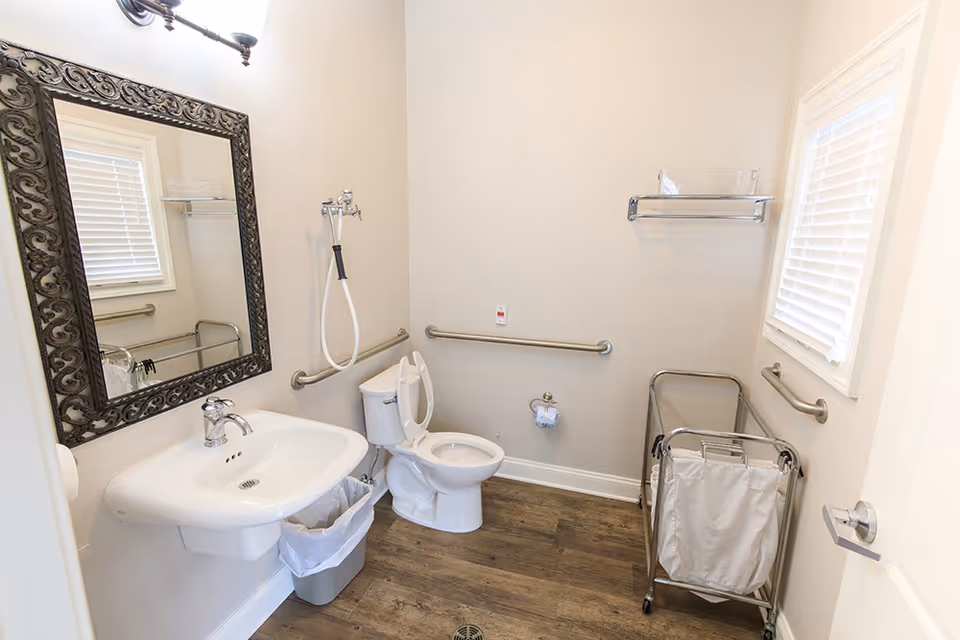 Accessible senior living bathroom with sink, toilet, grab bars, mirror, and a laundry cart.