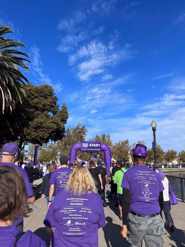 People in purple shirts walk outdoors toward a start arch on a sunny day.