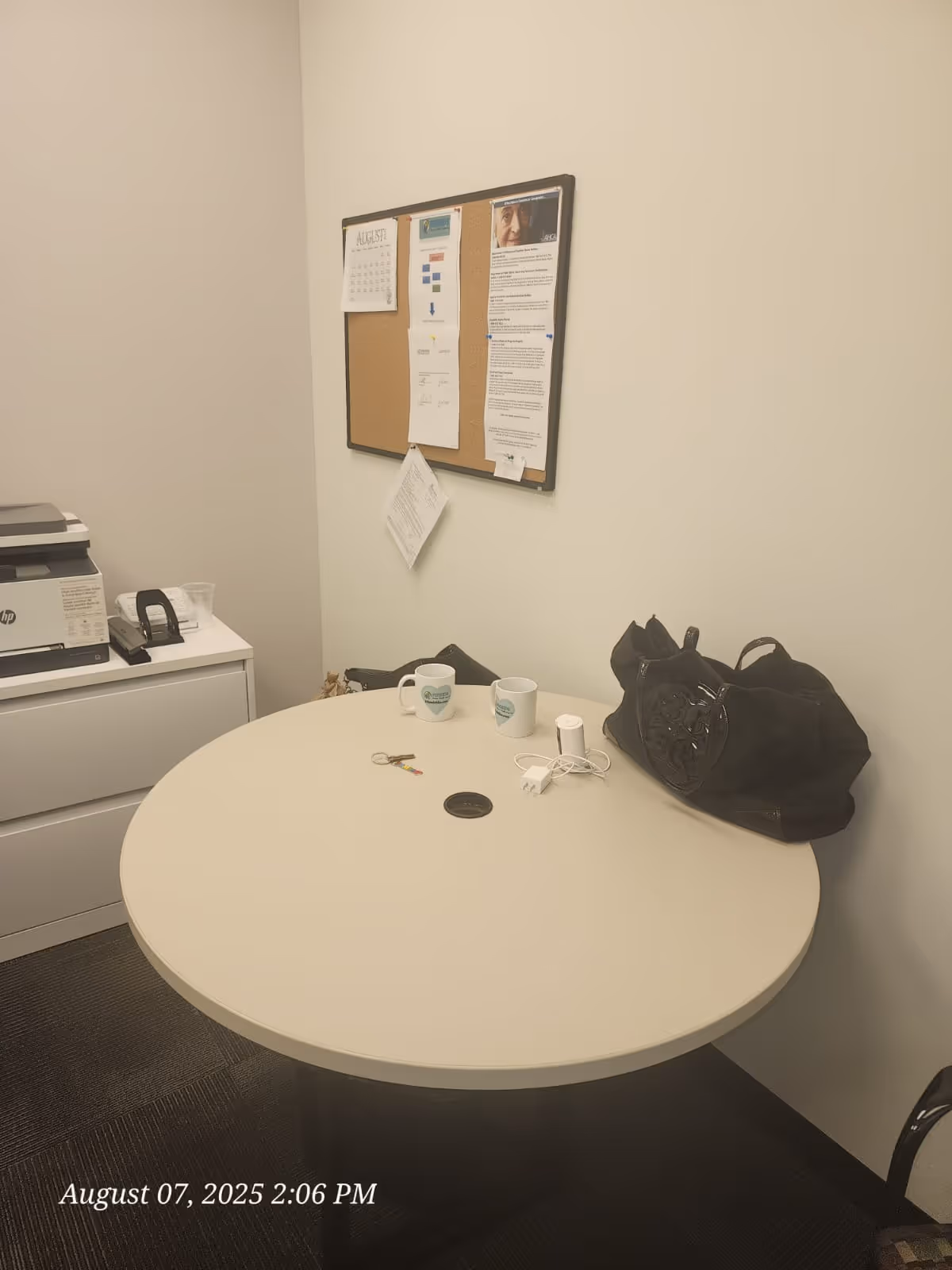 Round table with mugs, a bag, and a corkboard in a small office.