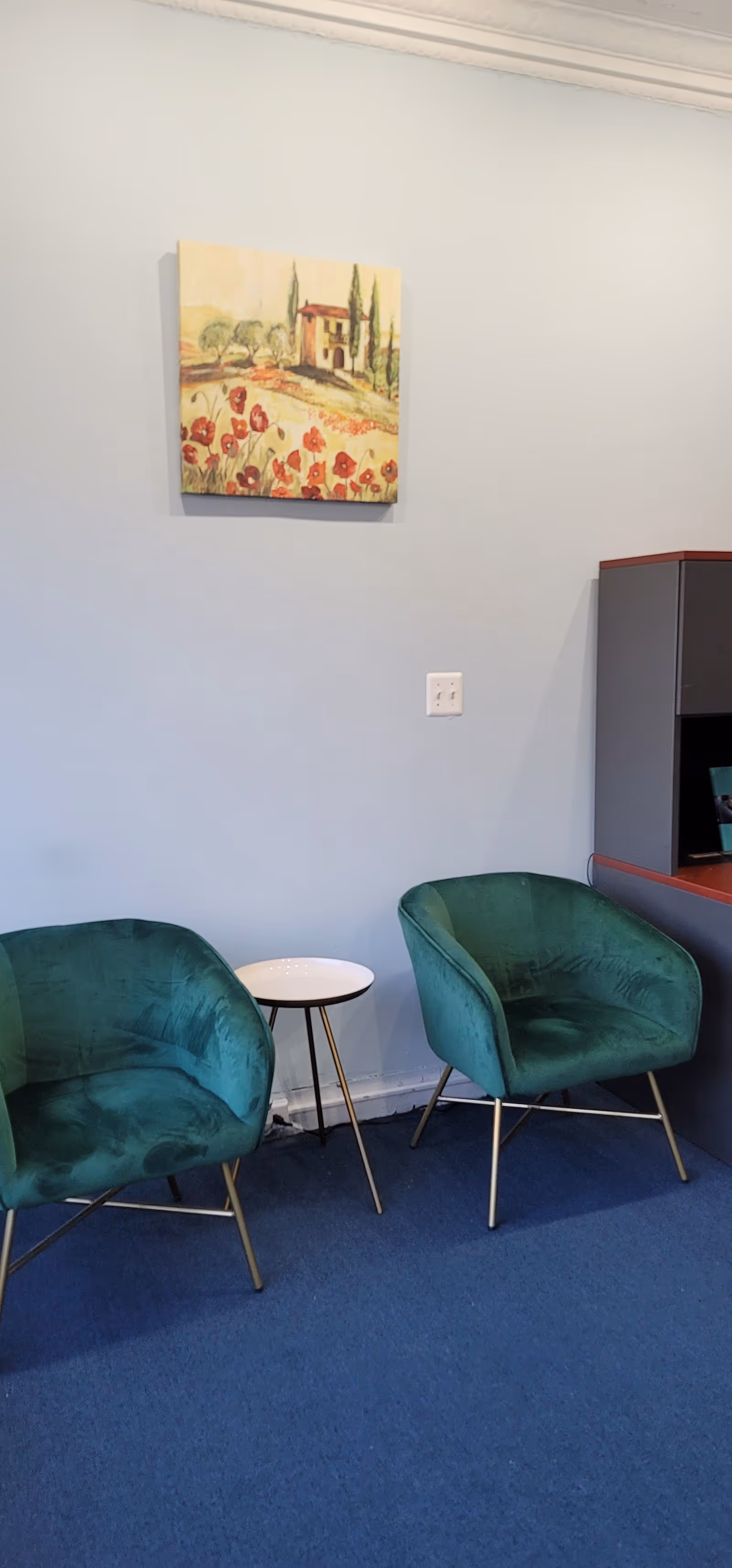 Two teal chairs and a small white table against a light blue wall with a painting