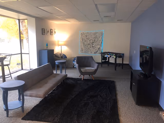 A cozy senior living lounge with sofa, chairs, rug, TV, foosball table, and a large window.