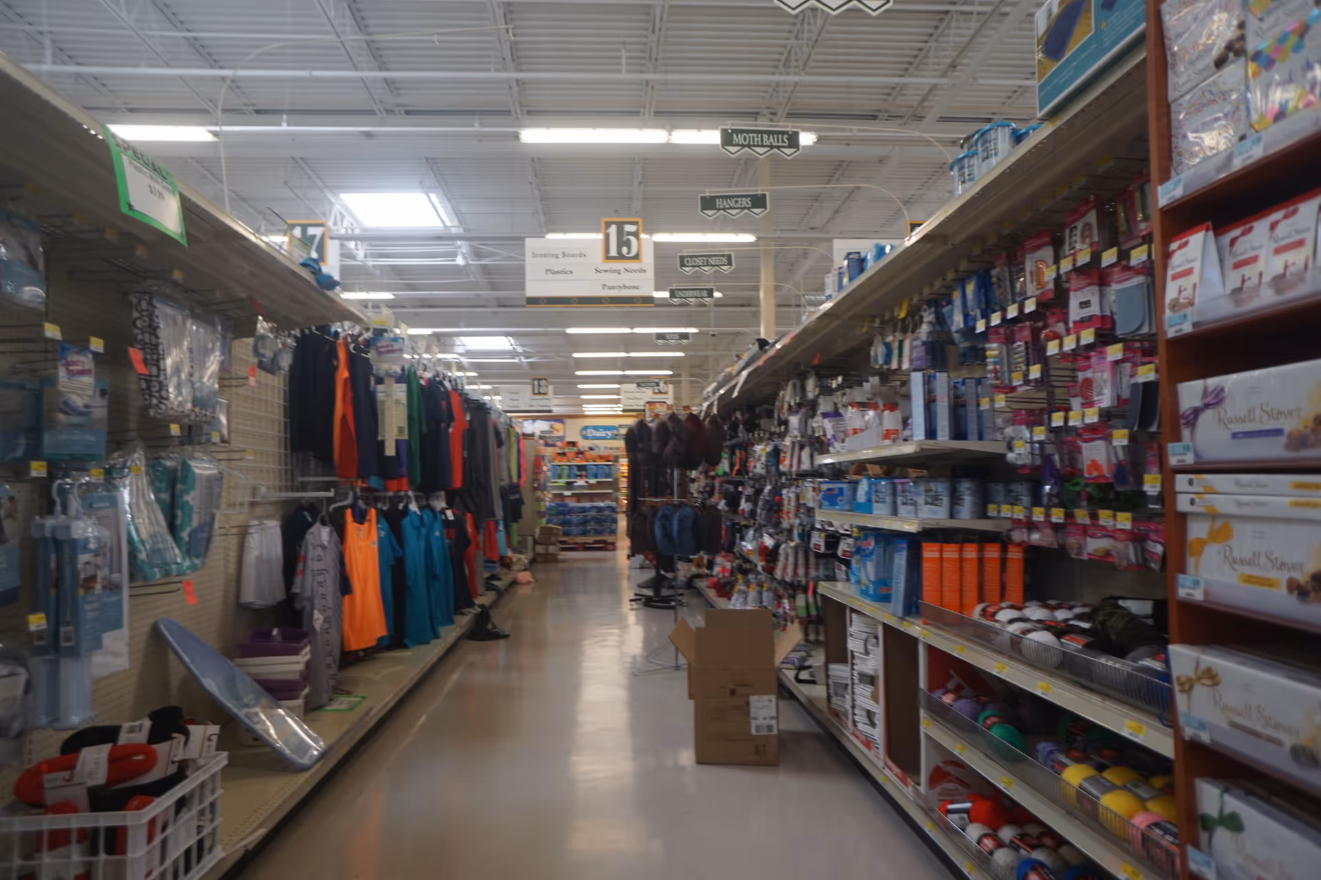 Long aisle inside Discount Drug Mart with clothes on left and shelves on right.