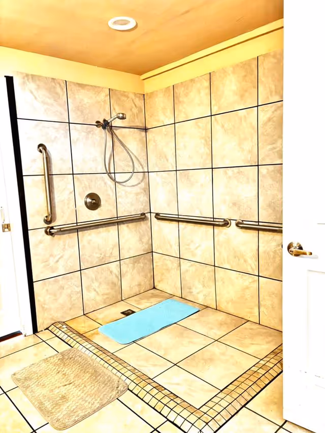 Accessible tiled bathroom with grab bars, a handheld shower, and two mats.