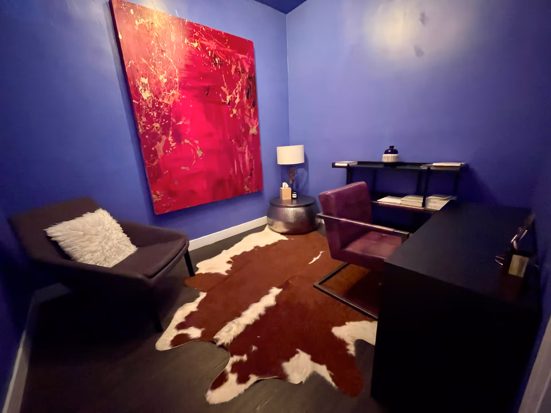 Blue-walled office-like room with a red abstract painting, desk, chair, and cowhide rug.