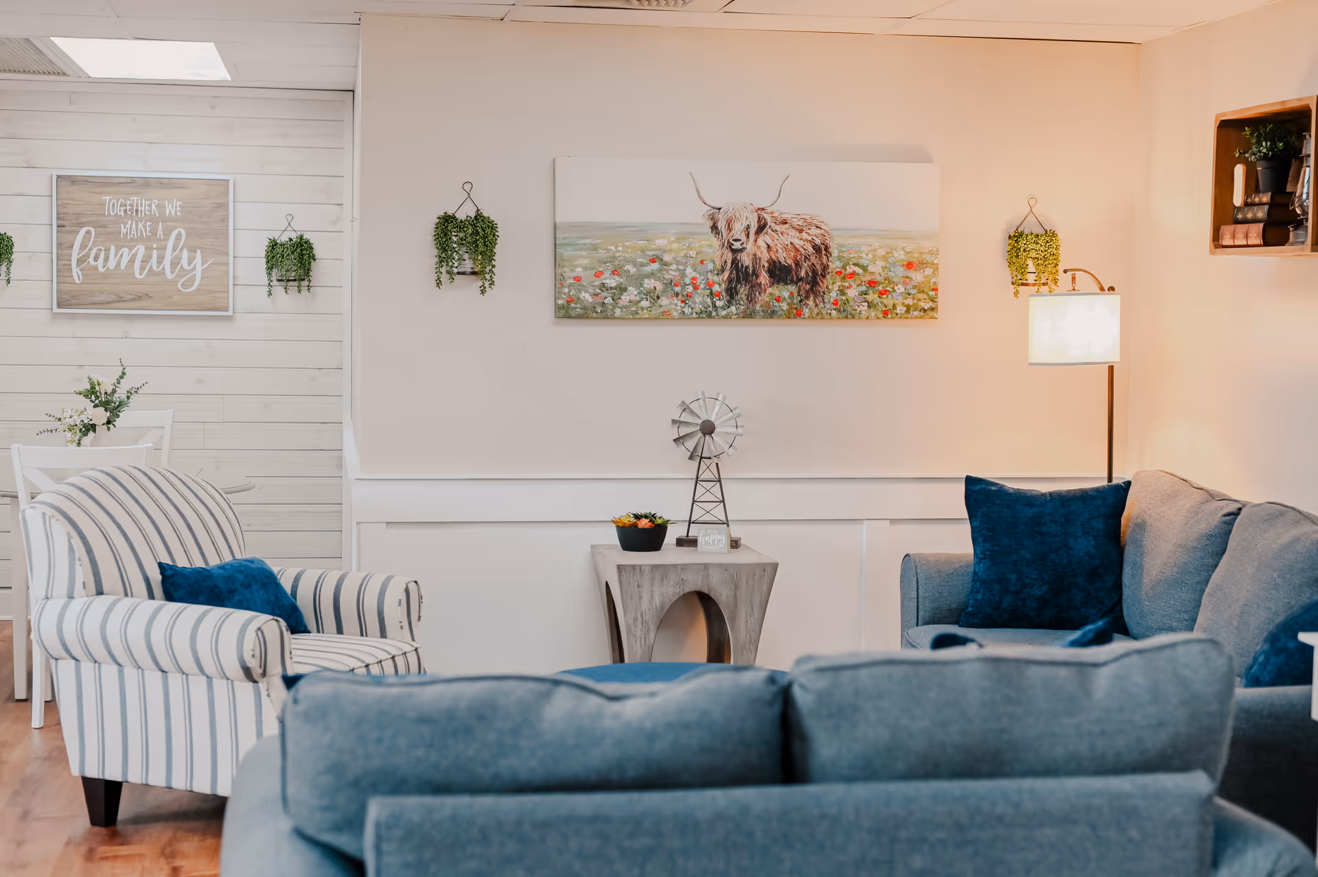 A bright living room in a senior care facility with blue seating, wall art, and plants.