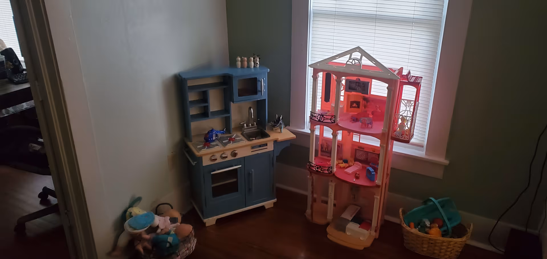 Blue toy kitchen and pink dollhouse sit by a window in a toy-filled room.