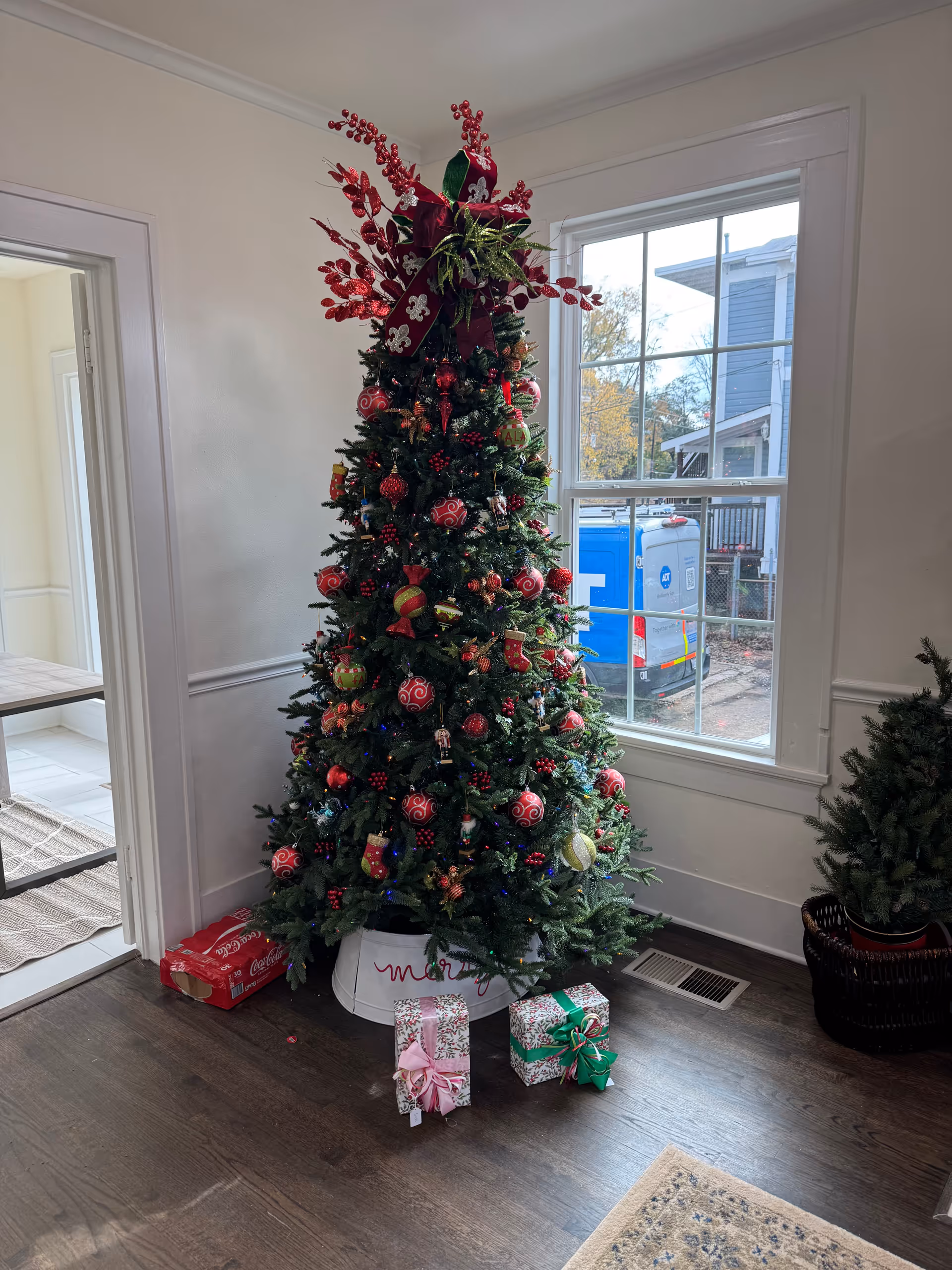 A Christmas tree with red ornaments and wrapped gifts indoors near a window.