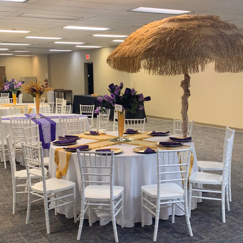 Decorated banquet tables with white chairs, purple napkins, and a thatched centerpiece indoors.