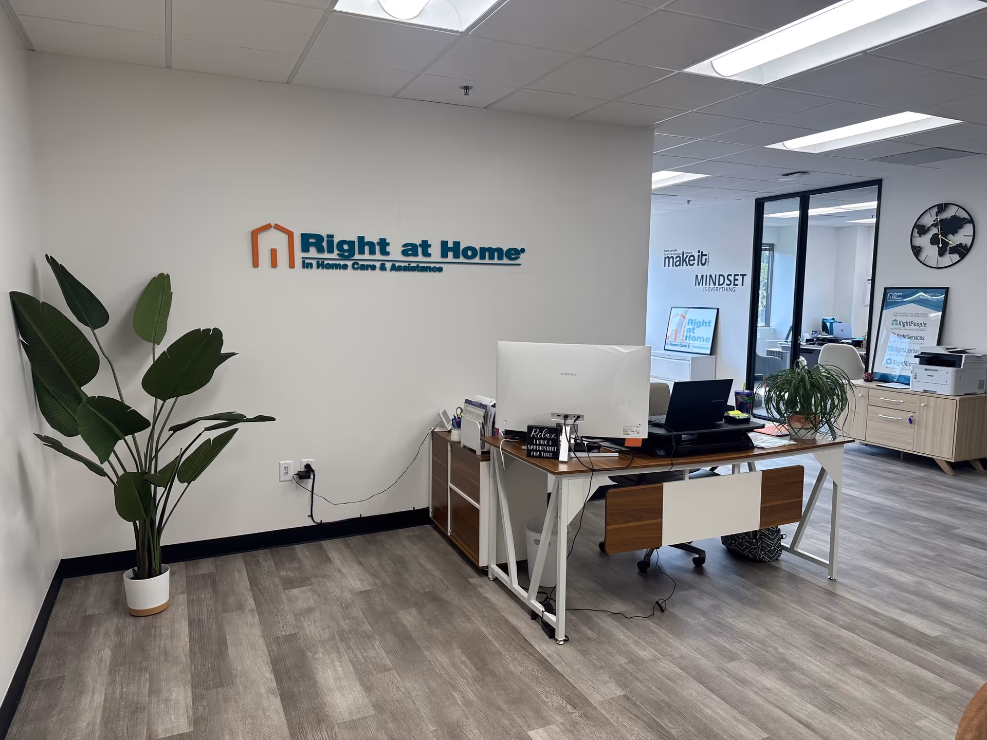 Reception area of Right at Home with desk, computer, plants, and logo on wall.