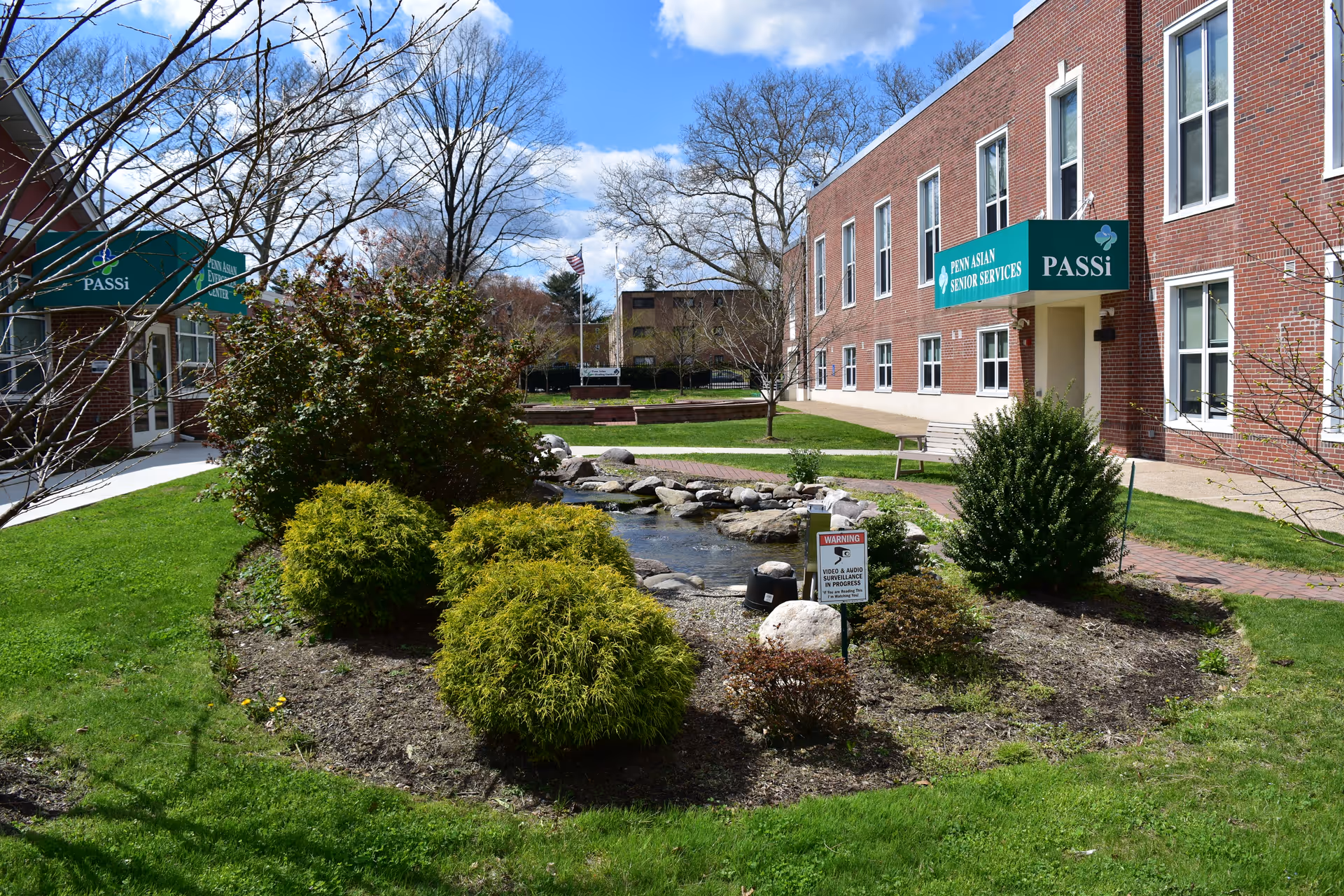 Brick PASSi facility with green signs, manicured garden, and small pond in foreground.