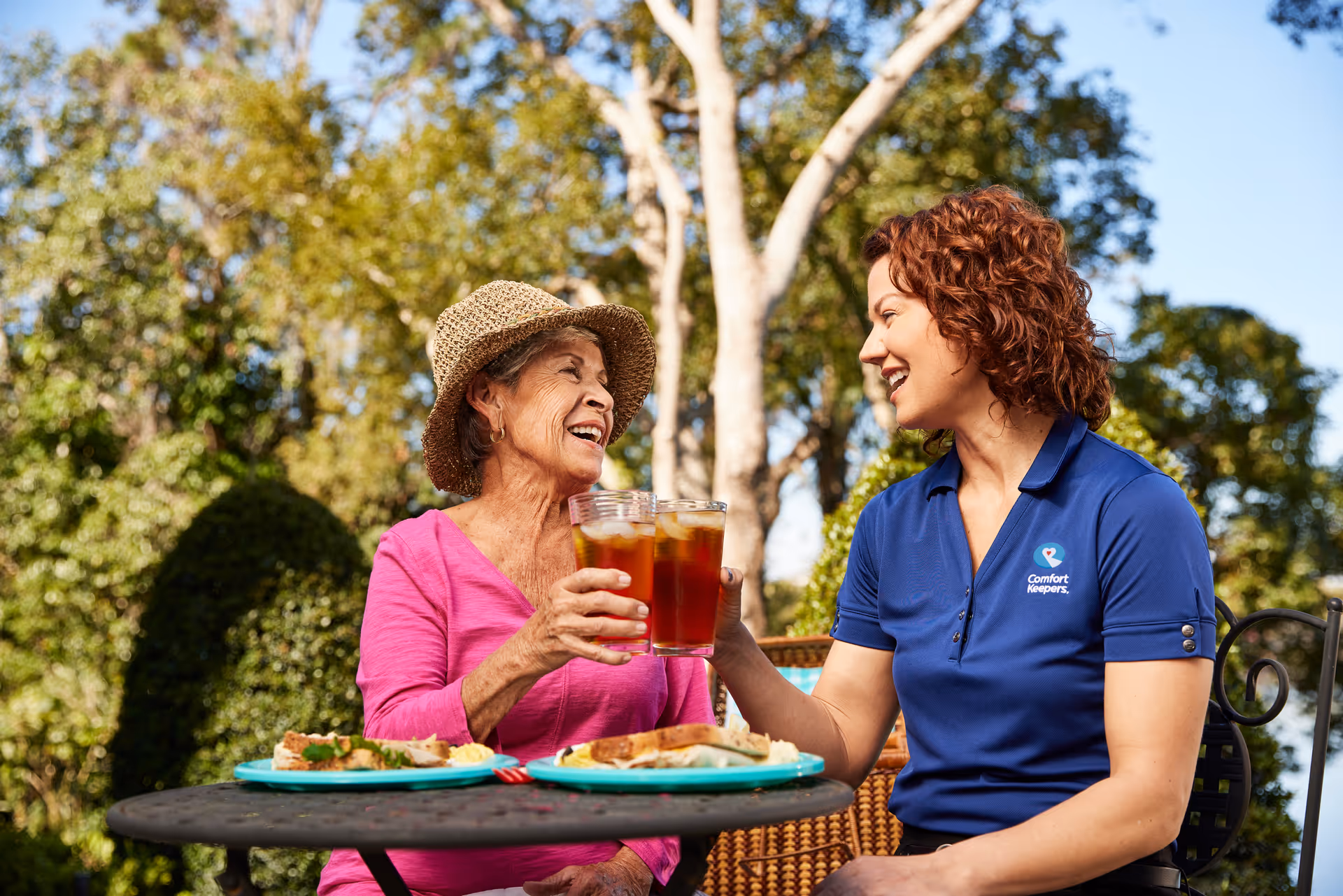 Senior resident and caregiver share drinks at an outdoor table with food.