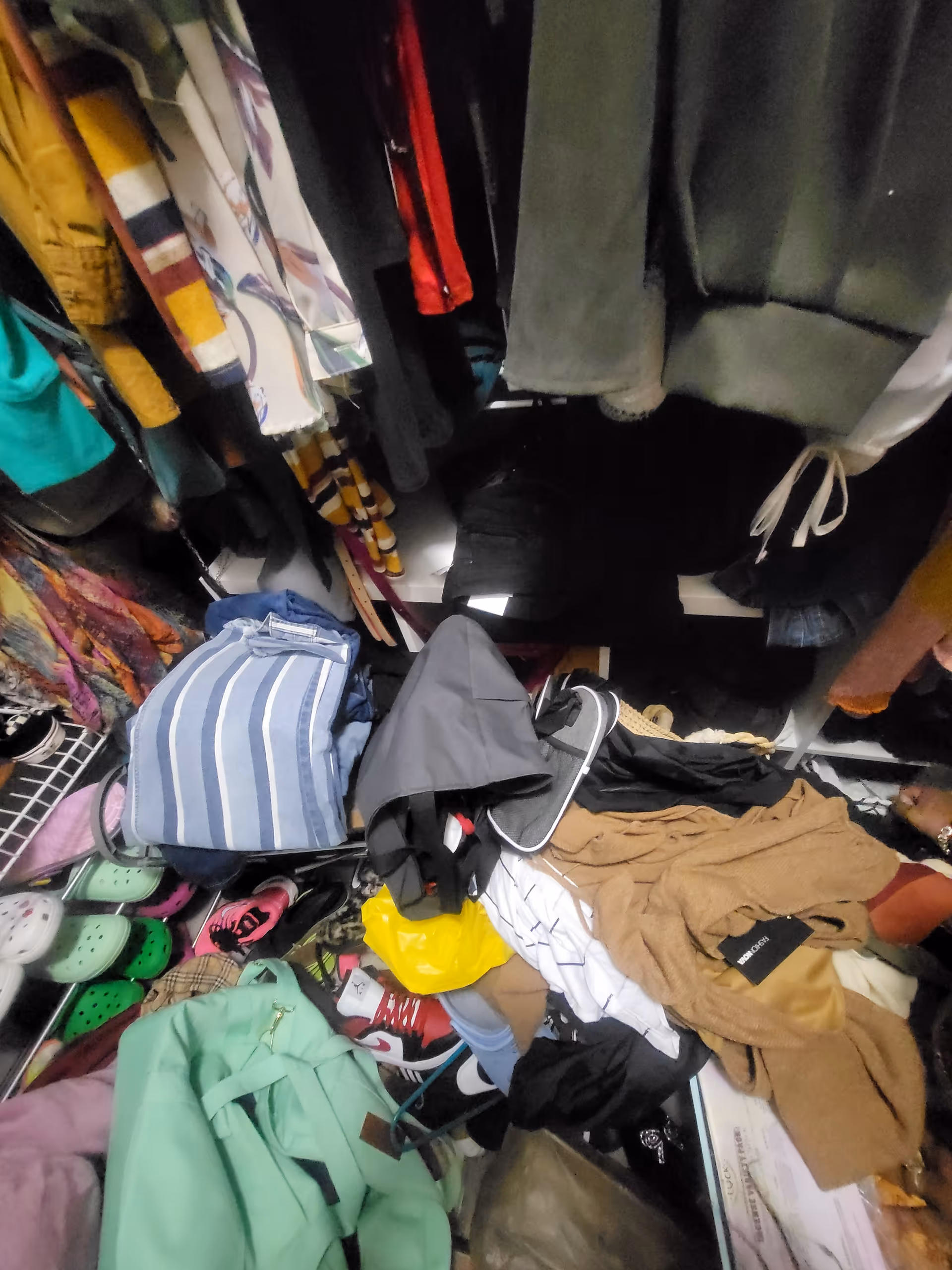 Cluttered indoor closet with hanging clothes, shoes, bags, and folded garments piled together.