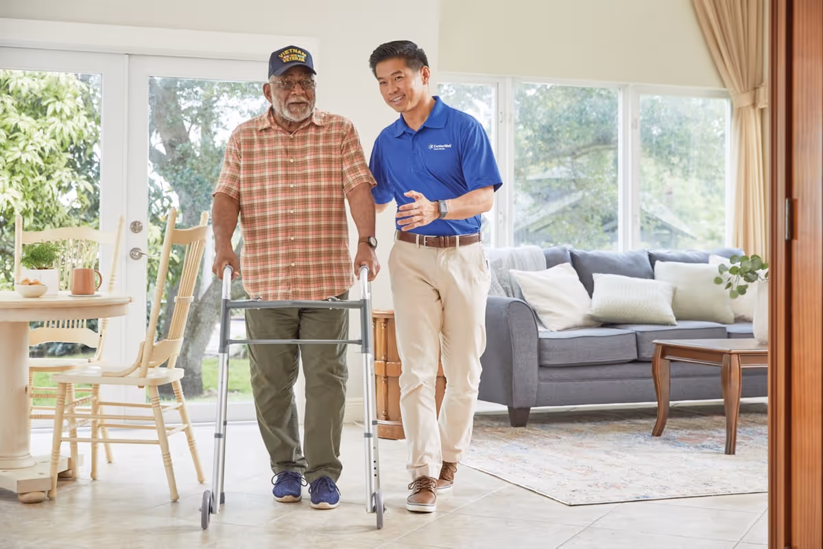 A caregiver assists an elderly man using a walker in a bright living room.