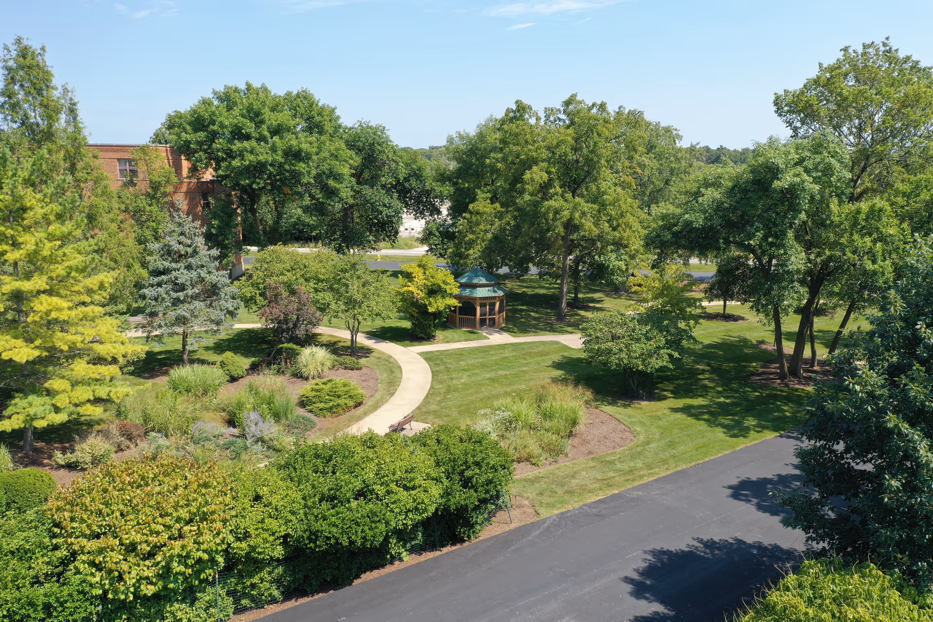 A sunny landscaped outdoor courtyard with a curved path and gazebo among trees.