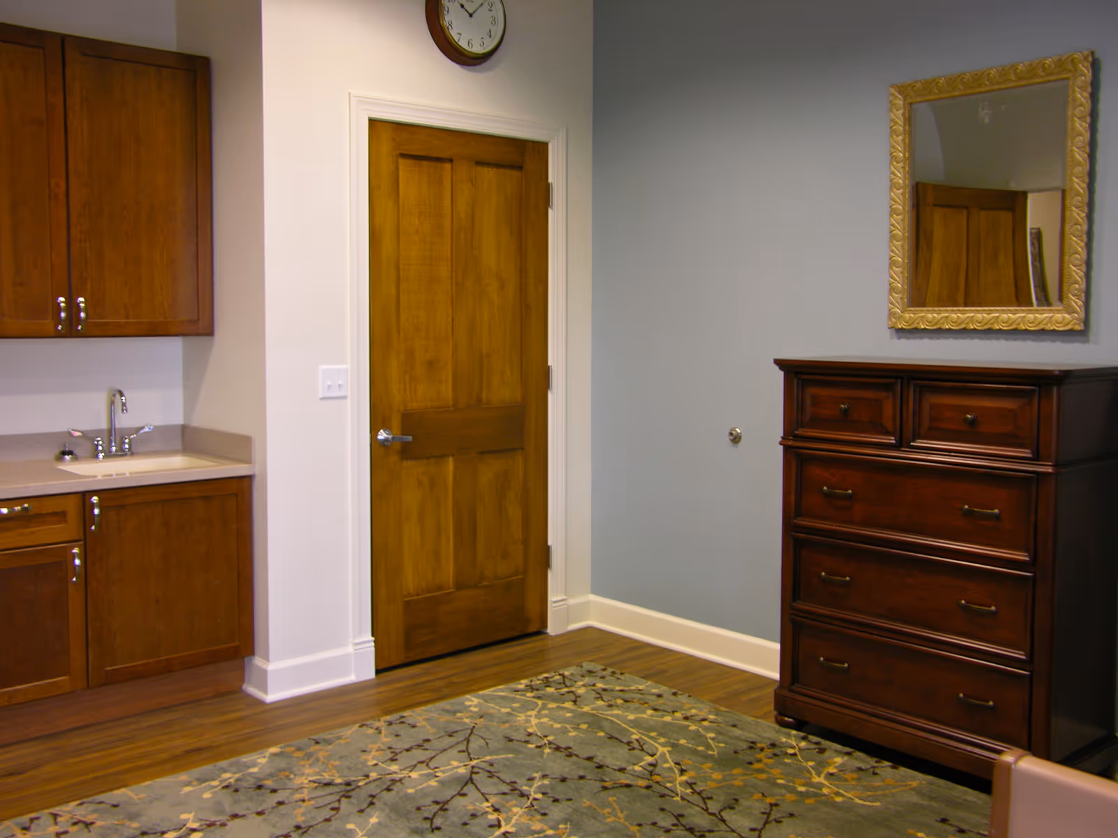 Interior of a senior living unit with wooden cabinets, a sink, door, dresser, and rug.