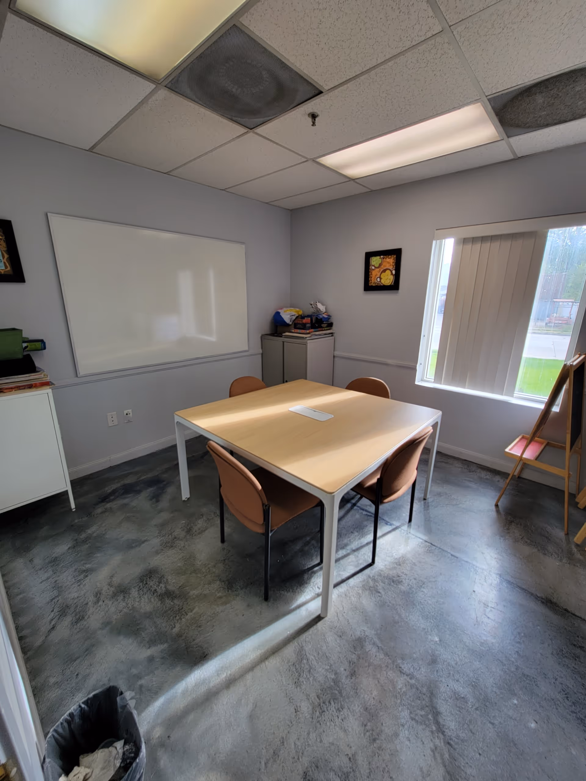 Small interior room with a rectangular table, four chairs, whiteboard, cabinets, and window blinds.